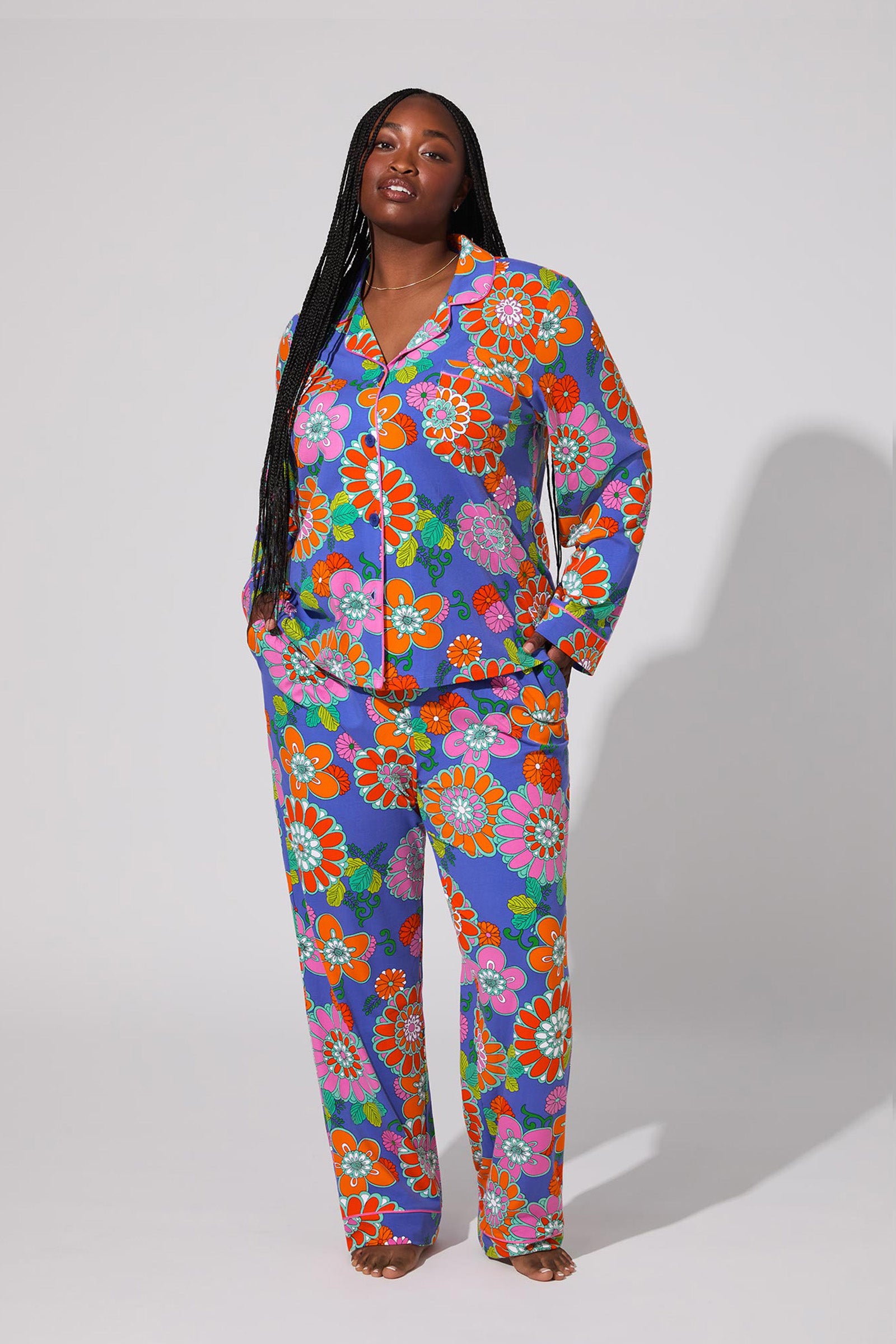 A woman with long braids stands barefoot against a light gray backdrop, confidently modeling the Classic PJ Set - Tokyo Floral, featuring a bold, colorful floral pattern on a purple background.