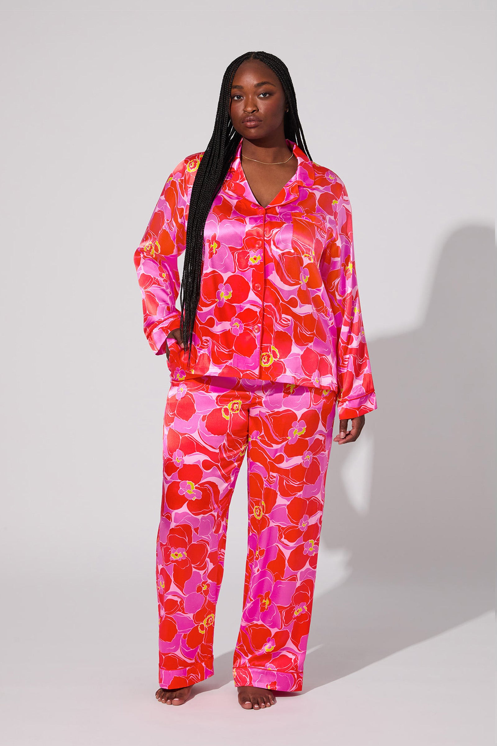 A woman stands barefoot in the Silk Classic PJ Set - Winter Orchids, featuring a vibrant red and pink floral pattern, long sleeves, and a button-up top, against a plain light background.