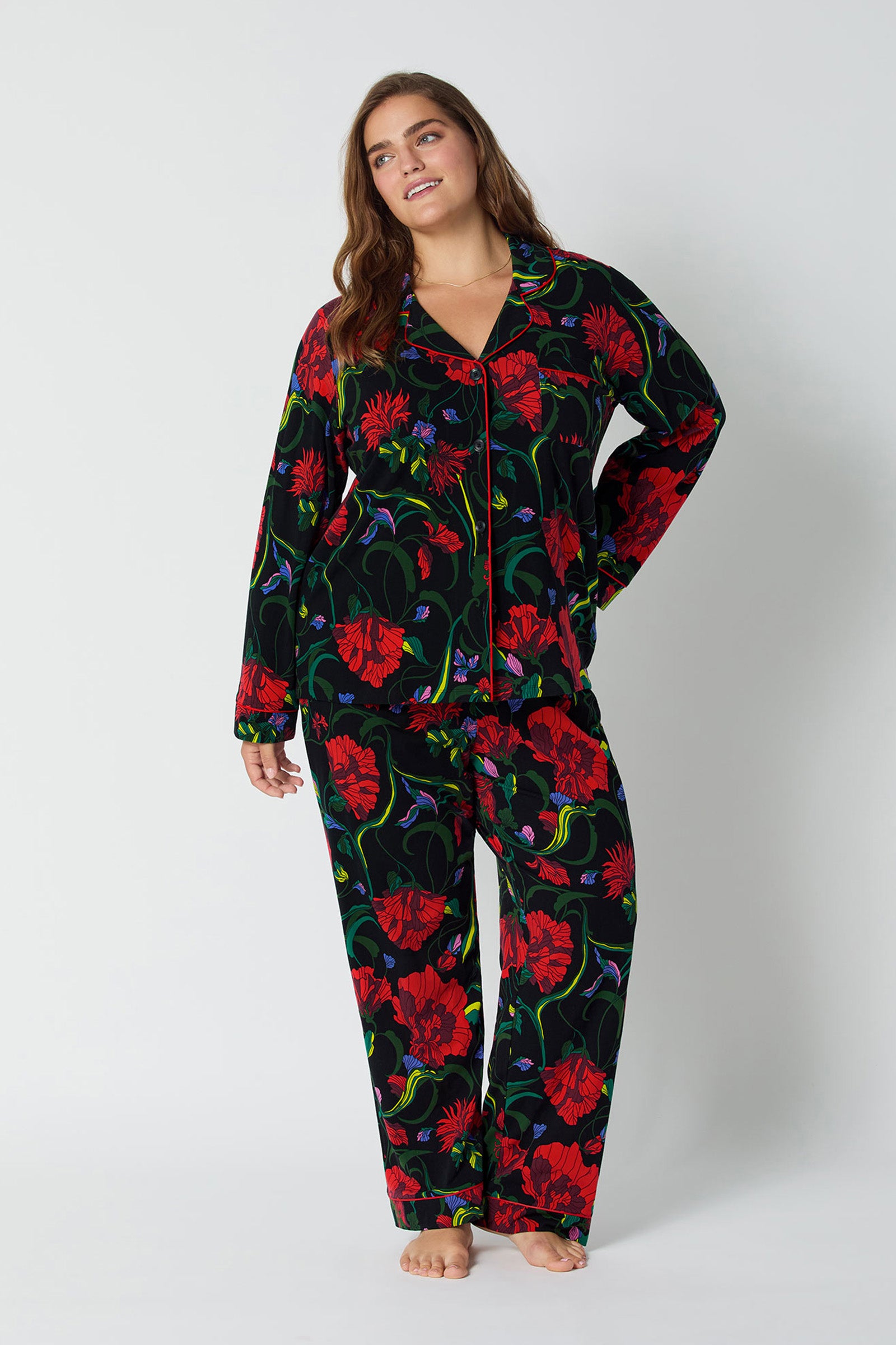 A woman stands barefoot against a plain background, wearing the Haute Floral L/S Classic Pj—a matching set with a black base and bold red floral print. The pajama top features long sleeves and a collar, paired with full-length pants.