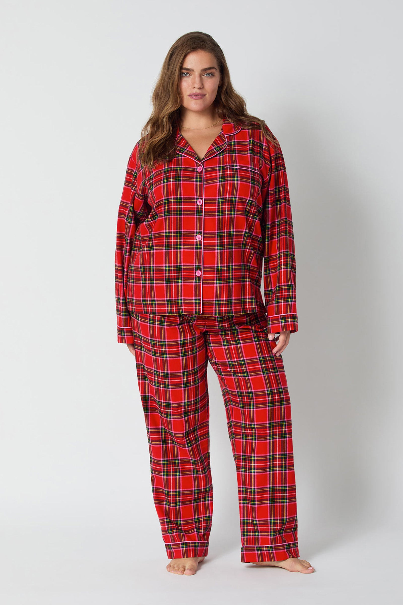 A woman with long brown hair stands barefoot against a plain background, wearing the Christmas Plaid L/S Classic Pj set featuring a matching red plaid button-up top and long pants.
