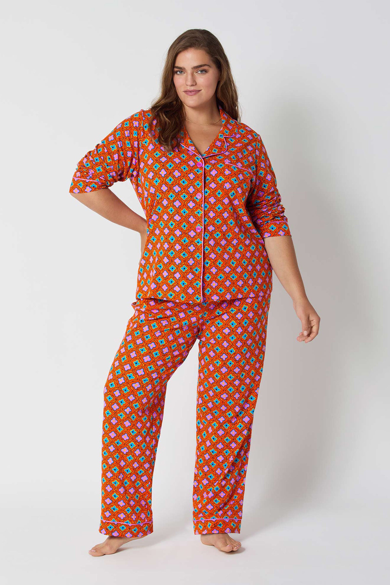 A woman stands barefoot against a white background, wearing the Daisy Foulard L/S Classic Pj—a matching long-sleeve pajama set with a red base and colorful geometric pattern, featuring a button-up collared top and loose-fitting pants.