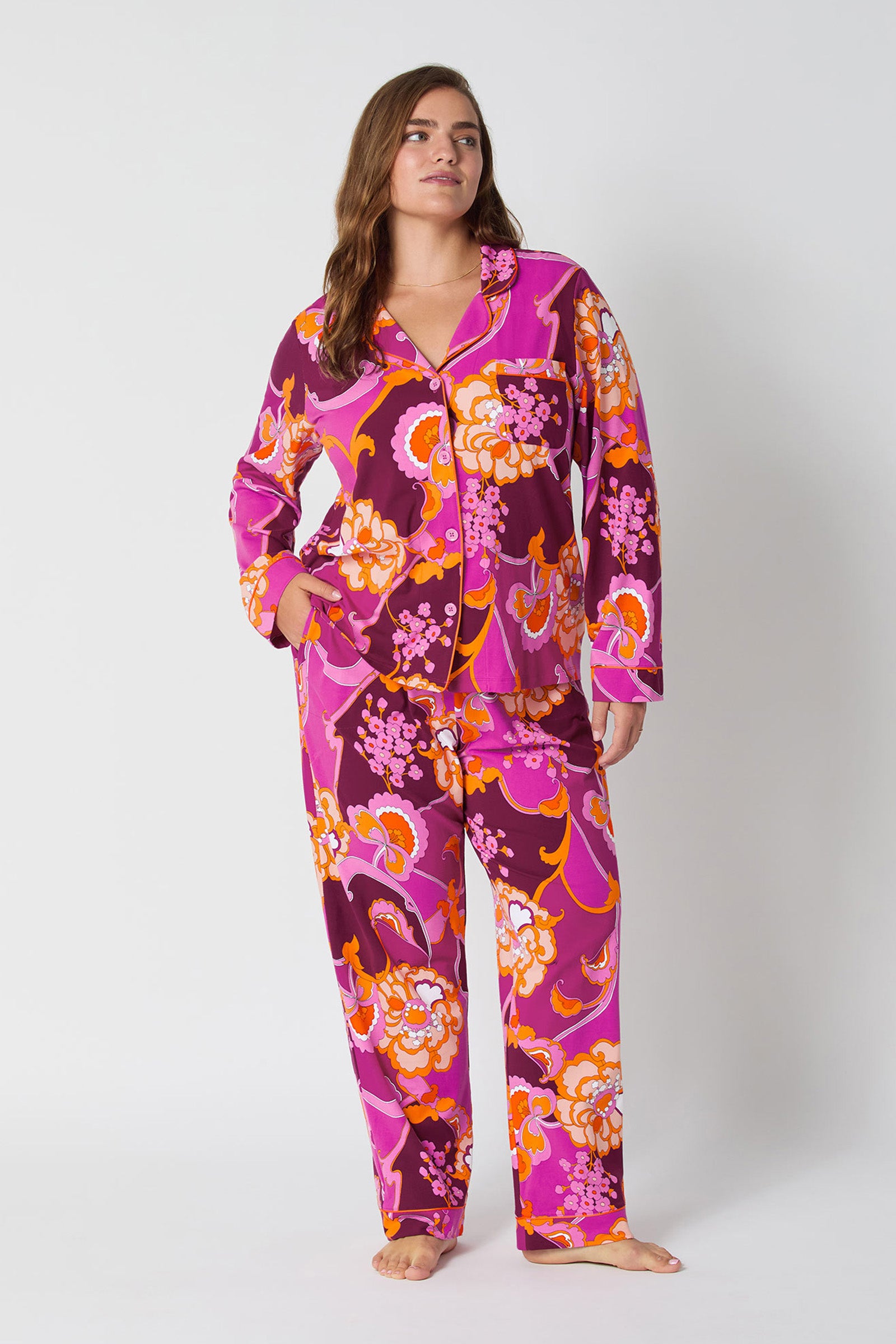 A woman with long hair stands barefoot against a white background, relaxed and confident, wearing the vibrant Apache Plume L/S Classic Pj featuring bold pink, orange, and purple floral patterns in a loose-fitting style.