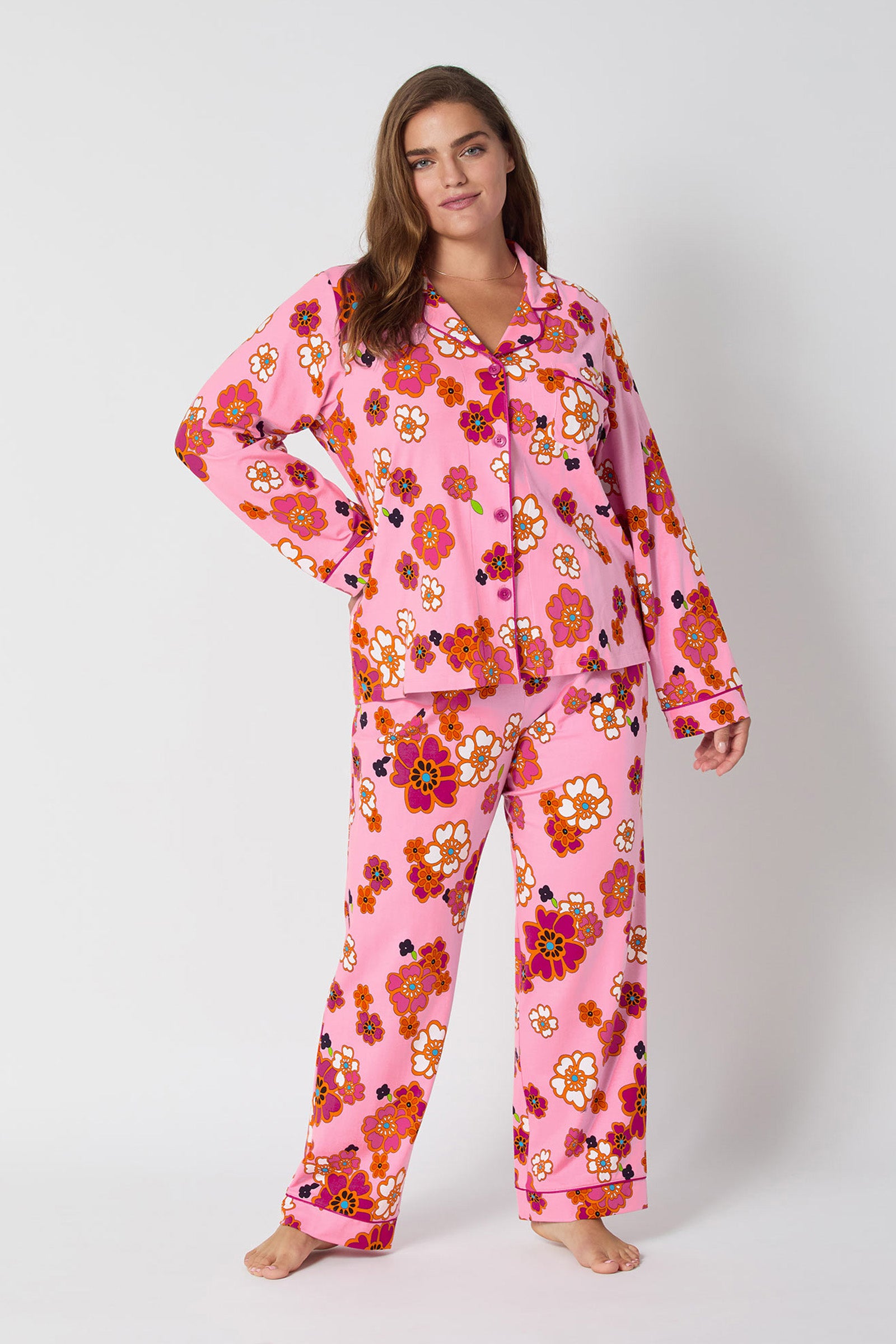 A woman stands barefoot against a plain background, wearing the Resort Floral L/S Classic Pj—pink pajamas with floral patterns, long sleeves, and pants. One hand rests on her hip as she displays a relaxed, confident expression.