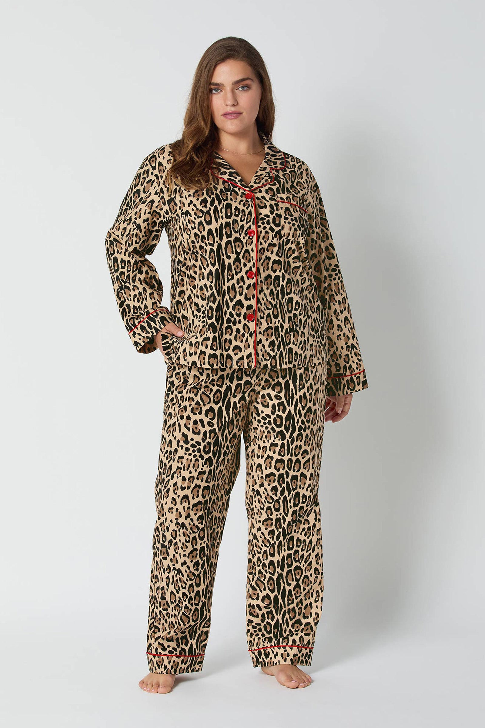 A woman with long brown hair stands barefoot against a white background, wearing the Leopard Stripe L/S Classic Pj set—long-sleeve top with red buttons and trim, matching leopard print pants, and a relaxed expression.