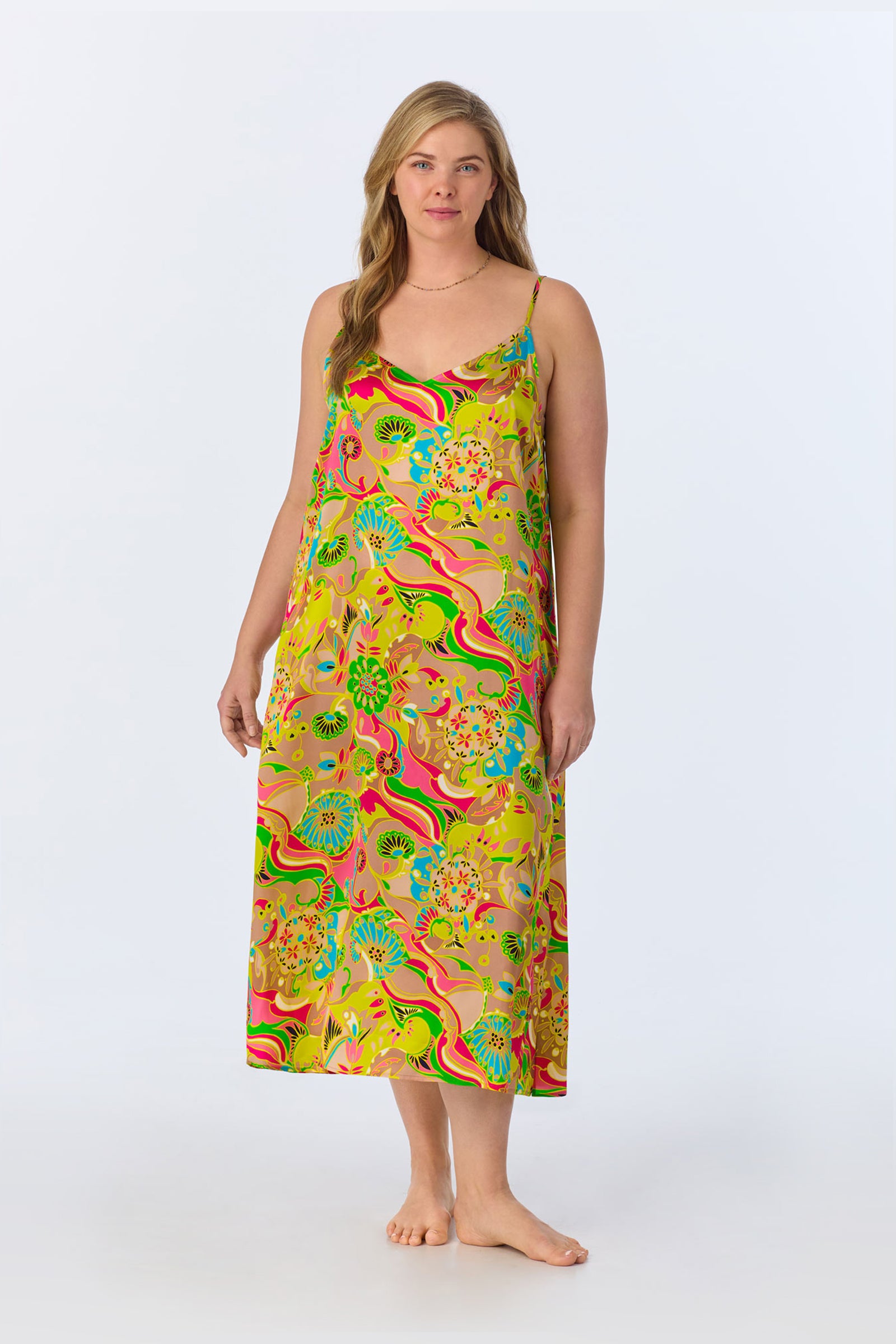 A woman with light hair and a neutral expression stands barefoot against a plain background, wearing the TT Maxi Chemise Nightgown - Heritage Silk, a long, sleeveless dress with thin straps and a vibrant multicolored pattern.