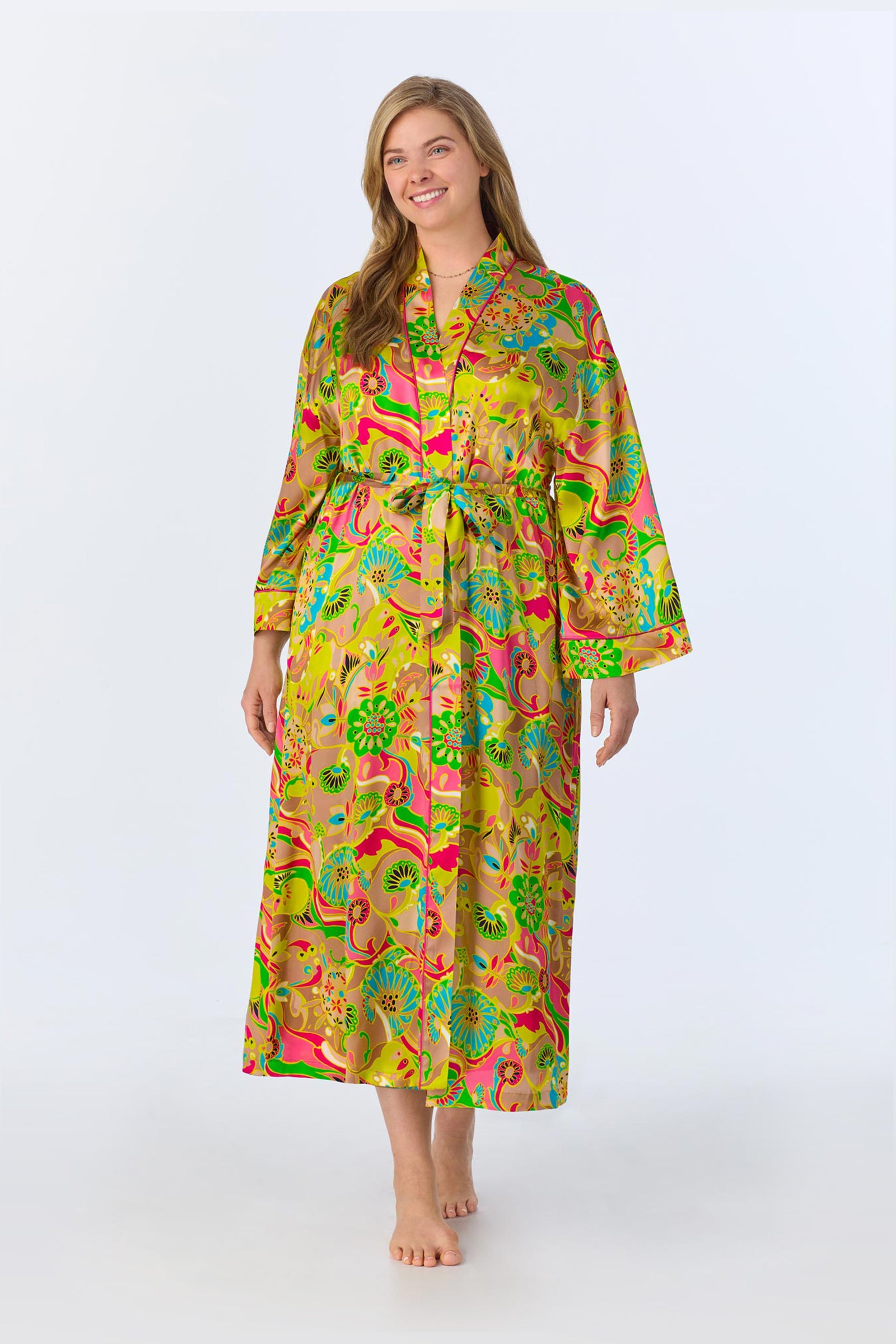 A smiling woman with long hair wears the TT Banded Collar Robe - Heritage Silk, a colorful, floor-length satin robe with vibrant floral and animal prints. She stands barefoot against a plain white background.