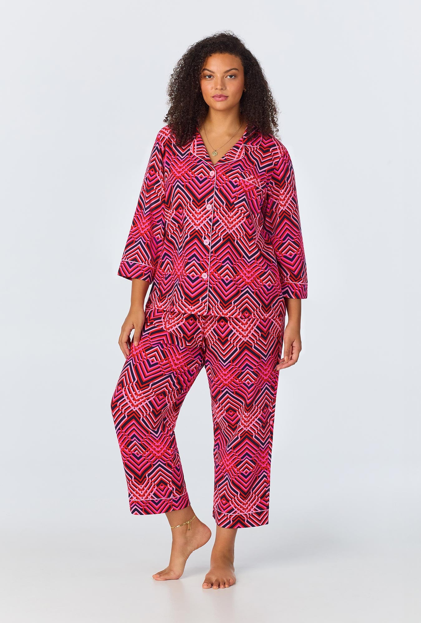 A woman stands barefoot in the Geo Trellis Womens 3/4-Sleeve Cropped Pant Jersey PJ Set, featuring a vibrant geometric pattern in red, pink, and black. This sleepwear includes a buttoned top with 3/4 sleeves and comfortable cropped pants.