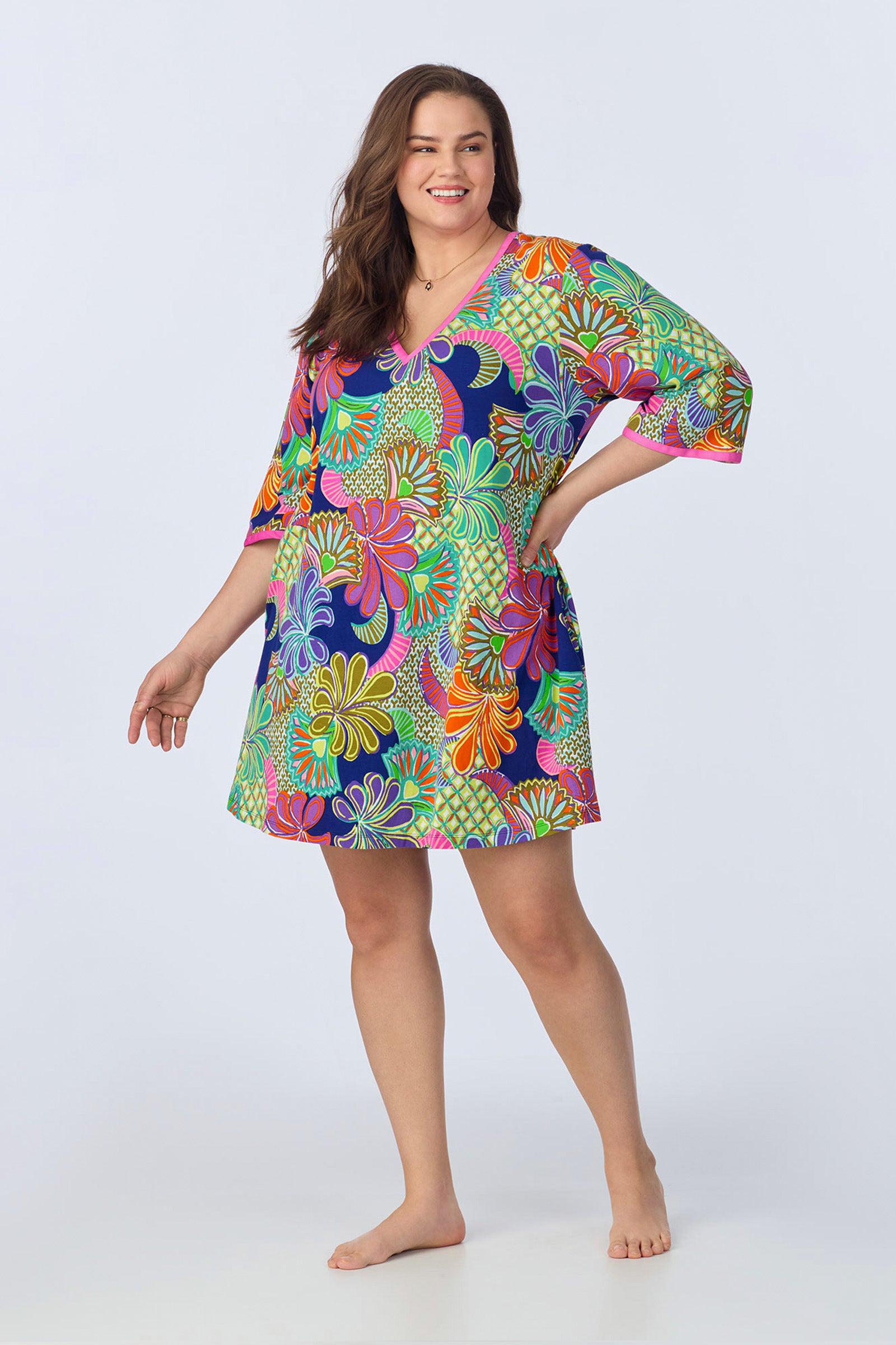 A smiling woman with long brown hair stands barefoot, wearing the PJ Caftan Dress - Zanzibar Stretch Jersey, a colorful floral short dress with three-quarter sleeves and a V-neck, against a plain light background.