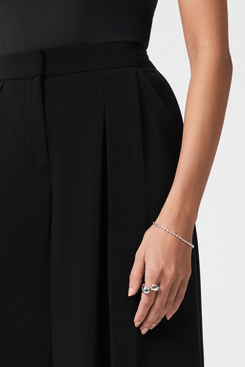 A woman wearing Saige Wide-Leg Pants is shown from the shoulders to mid-thigh. She has a delicate bracelet on her wrist and two rings on her fingers, with her right arm relaxed by her side against a plain background.