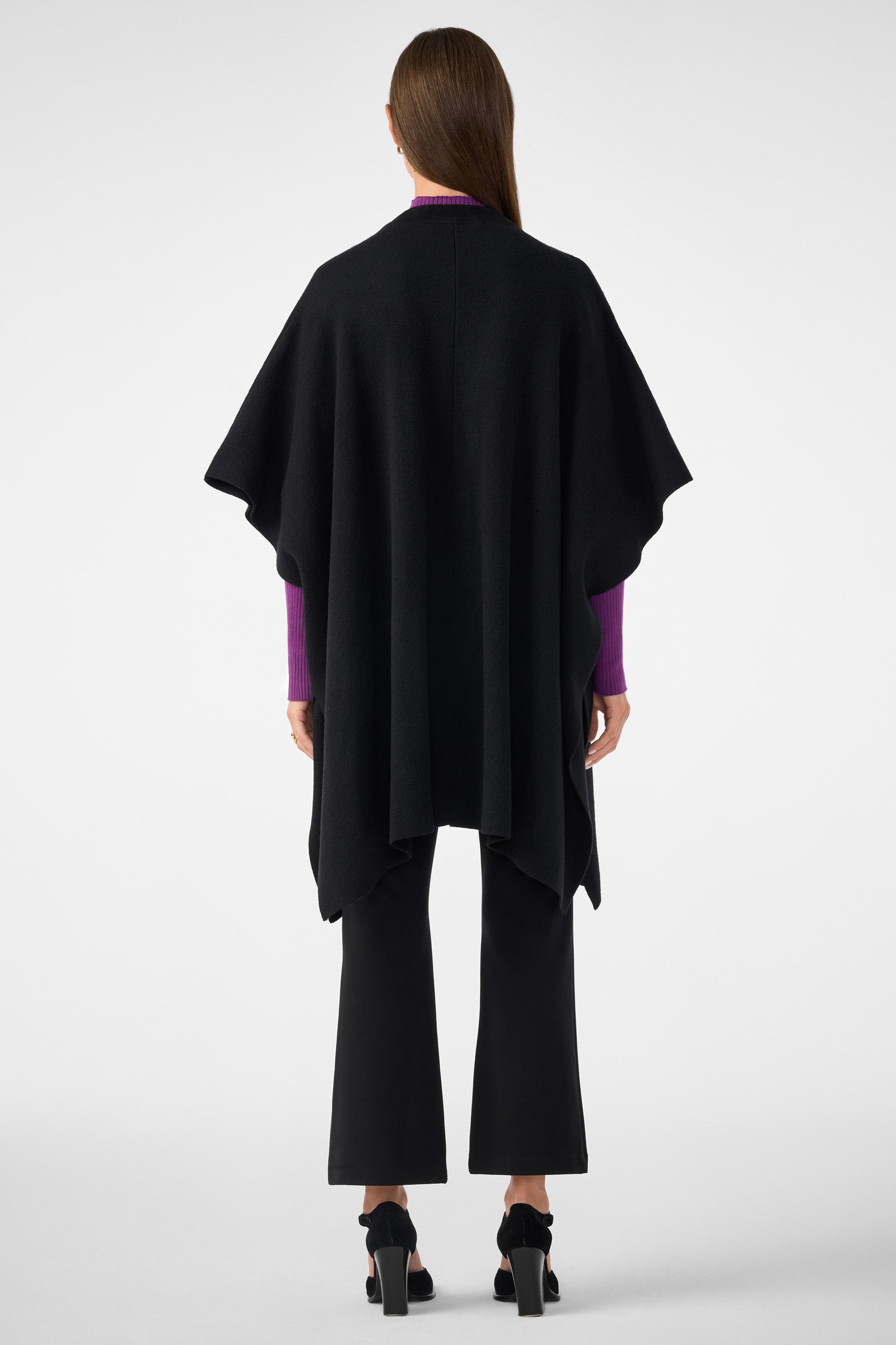 A woman faces away, wearing the Sapphire Poncho over a purple long-sleeve top, black pants, and black heeled sandals against a plain white background.