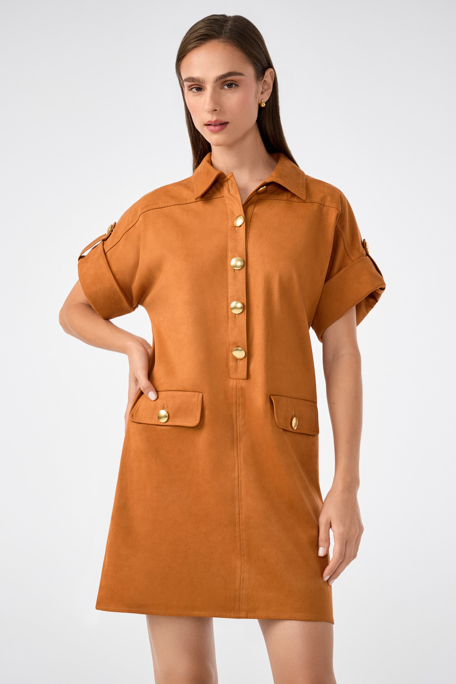 A woman stands against a plain light background wearing the Sequoia Dress—a short, brown button-up style with rolled sleeves, gold buttons, and two front flap pockets accented with gold buttons.