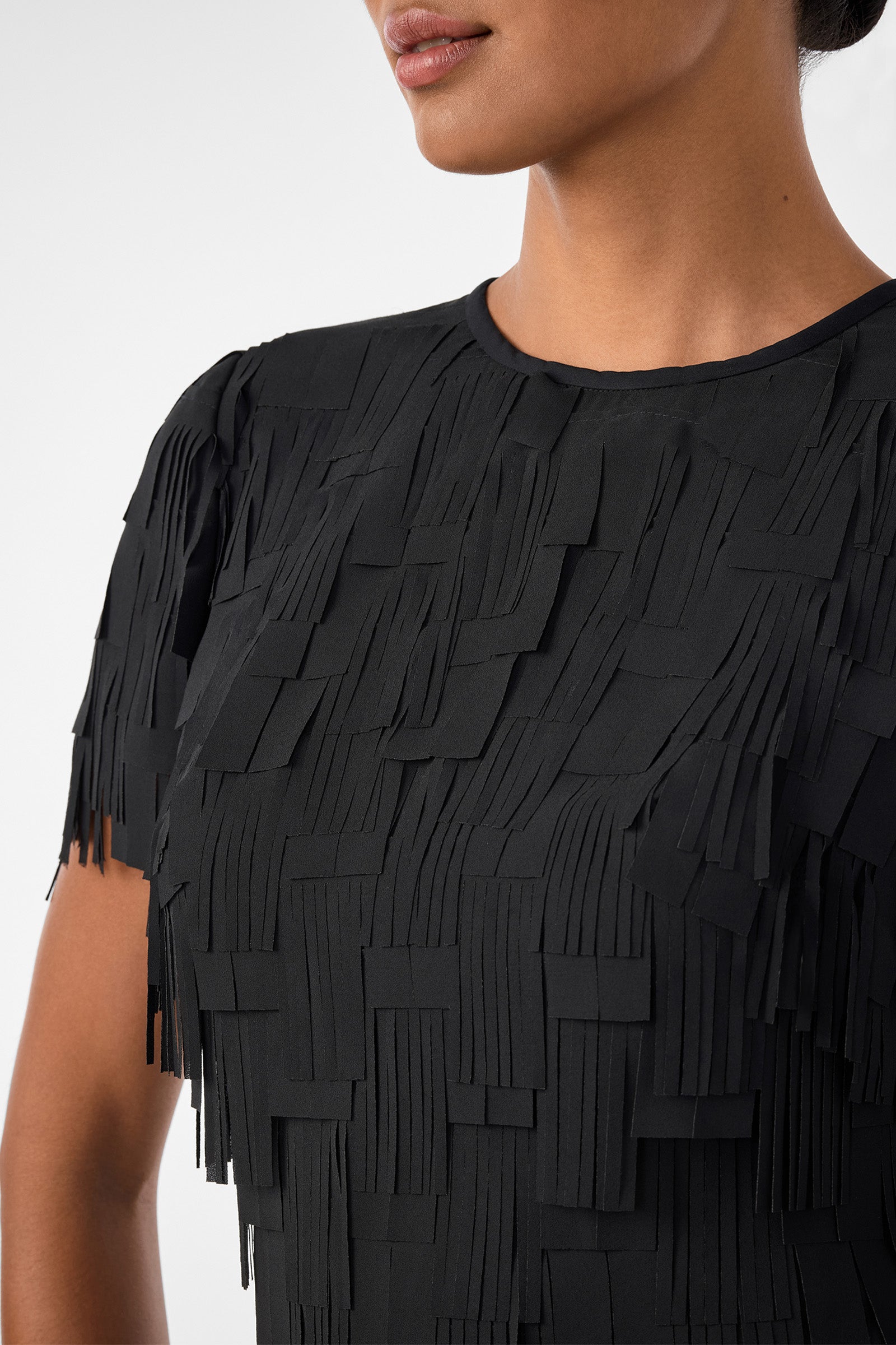 A woman wearing the Silkoak Dress, a black short-sleeved top with a textured pattern of layered, rectangular fringes. Only her lower face, neck, and upper torso are visible.