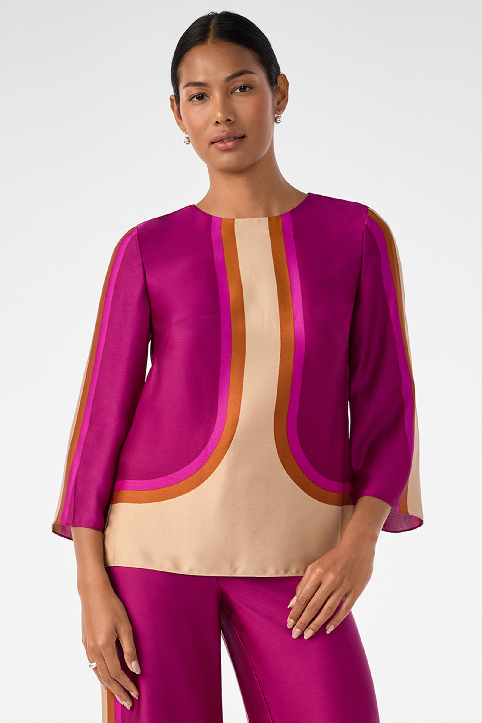 A woman stands facing forward in the Sprig 3 Top, a vibrant magenta blouse with orange and beige curved stripes and three-quarter sleeves. Her dark hair is pulled back, and she pairs the top with matching magenta pants.