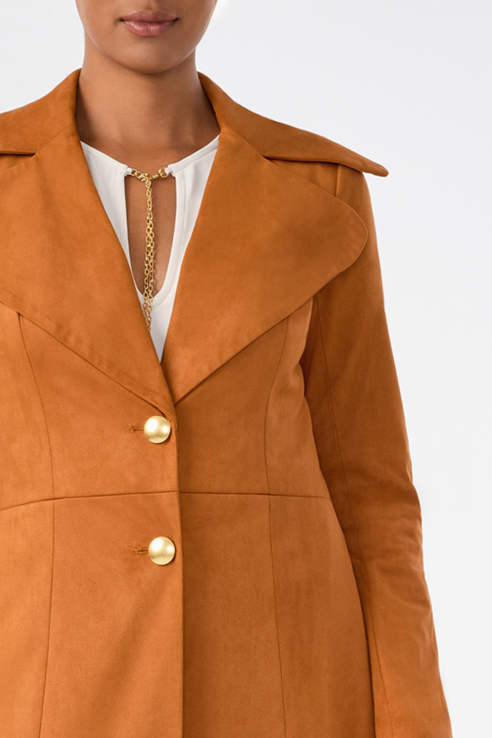 A person wears the Starflower Coat—a brown suede coat with large lapels and gold buttons—over a white blouse with a gold chain detail at the neckline. The image is cropped from shoulders to mid-thigh.