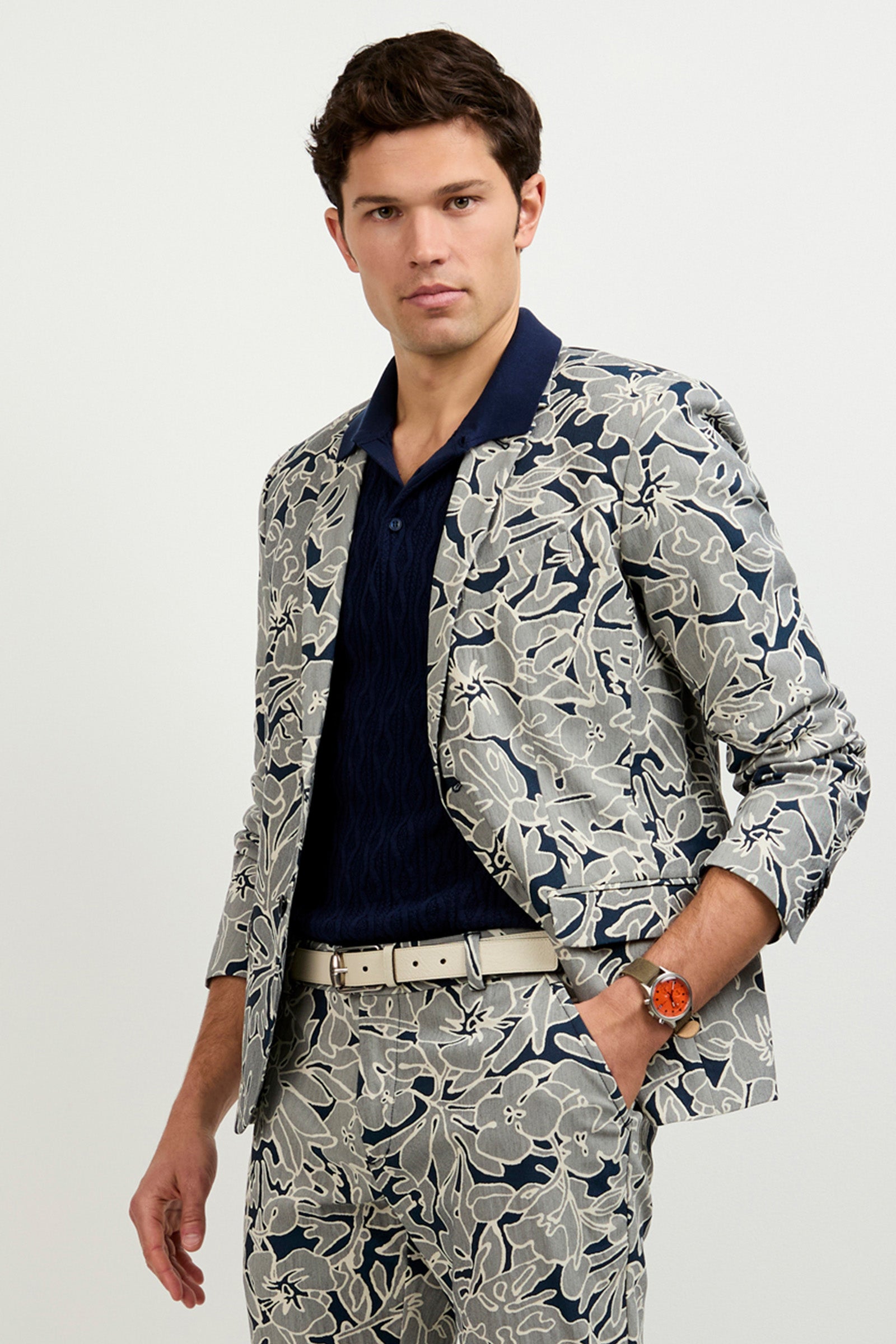 A man wearing the Thurston Blazer in artsy floral jacquard, paired with a navy textured shirt, light belt, and orange wristwatch, stands against a plain light background.