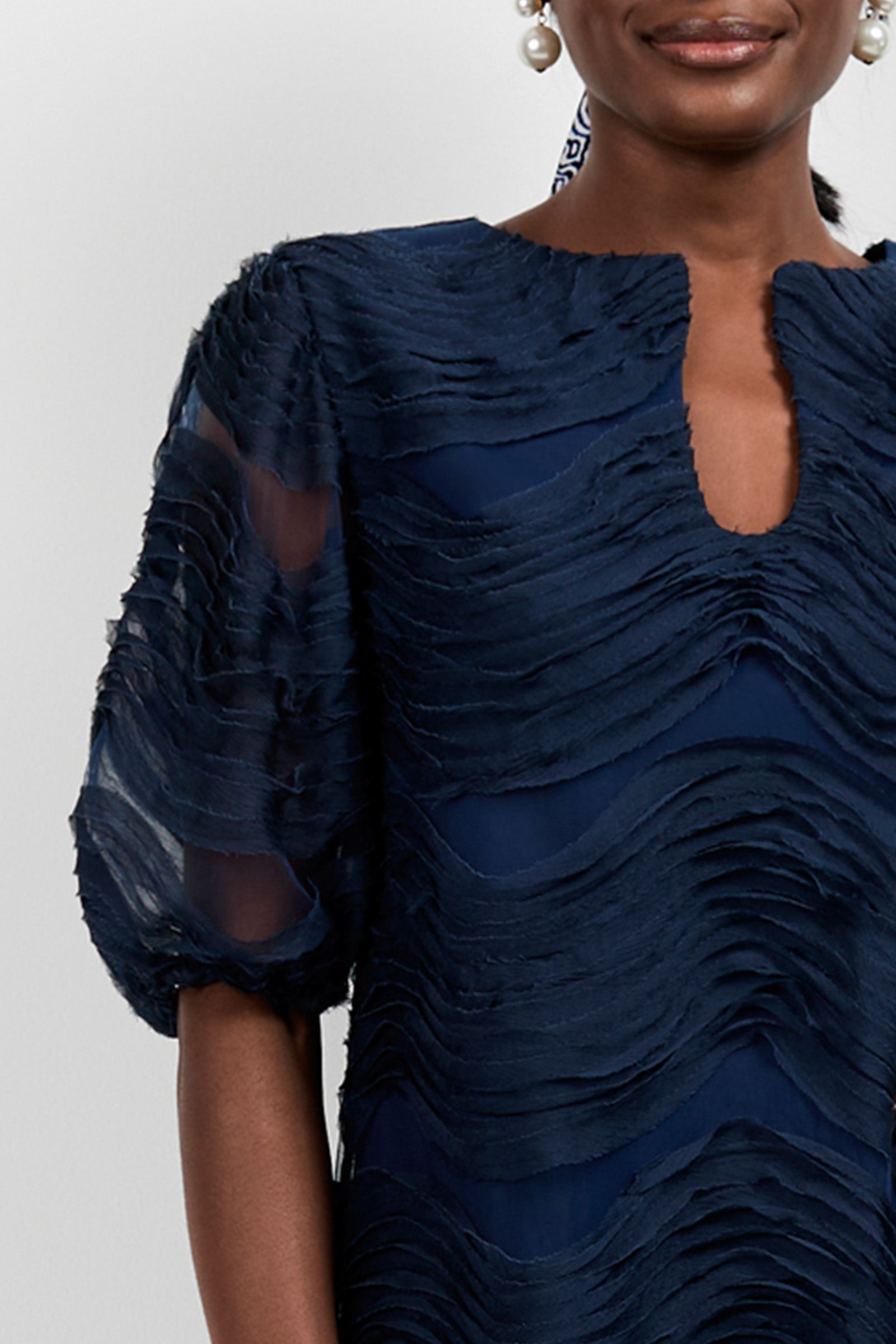 A woman wears the Torchino Organza Mini Dress, a navy blue textured piece with short puff sleeves and a round neckline with a keyhole opening; she has pearl earrings and only the lower half of her face is visible.