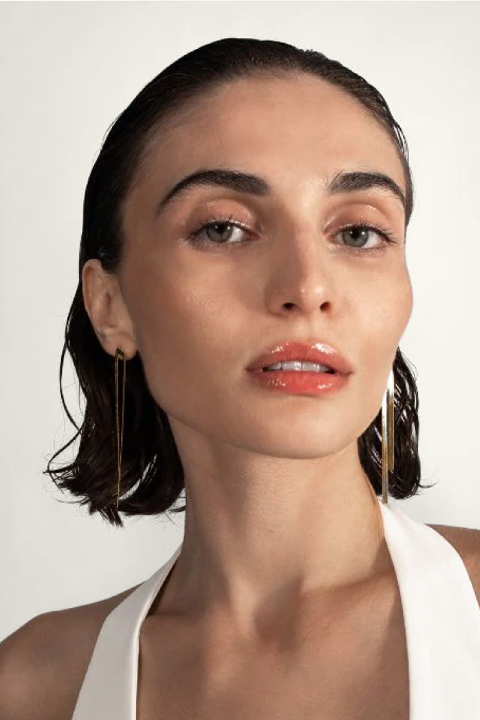 A woman with slicked-back dark hair and a white halter top confidently models the Lucien Earrings against a plain light background.