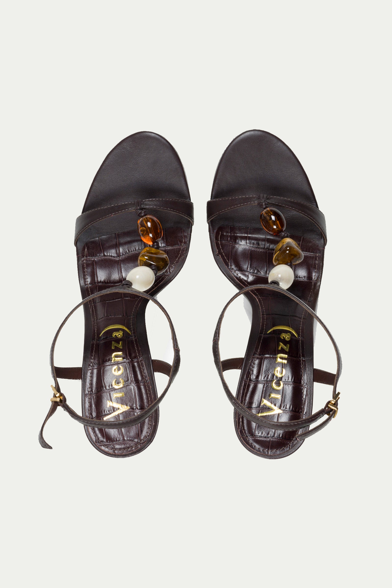 The Bloome Wedge Heel is a pair of dark brown open-toe sandals with ankle straps, featuring large amber, brown, and cream beads on the straps. The textured insoles display the VICENZA brand name in gold lettering.