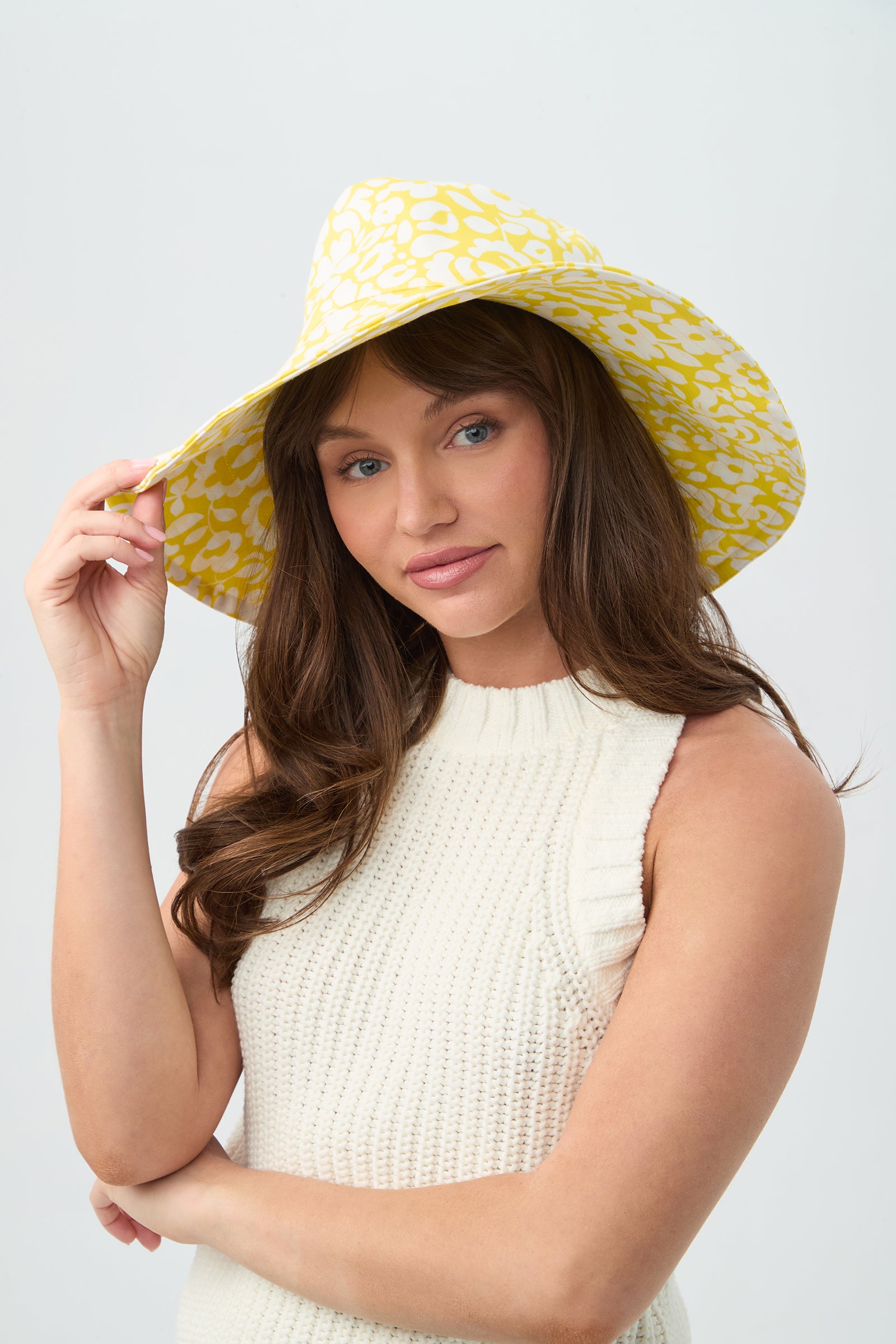 A woman with long brown hair wears the Shade Packable Sun Hat - Poolside Gossip and a sleeveless white knit top, looking at the camera against a plain light background.