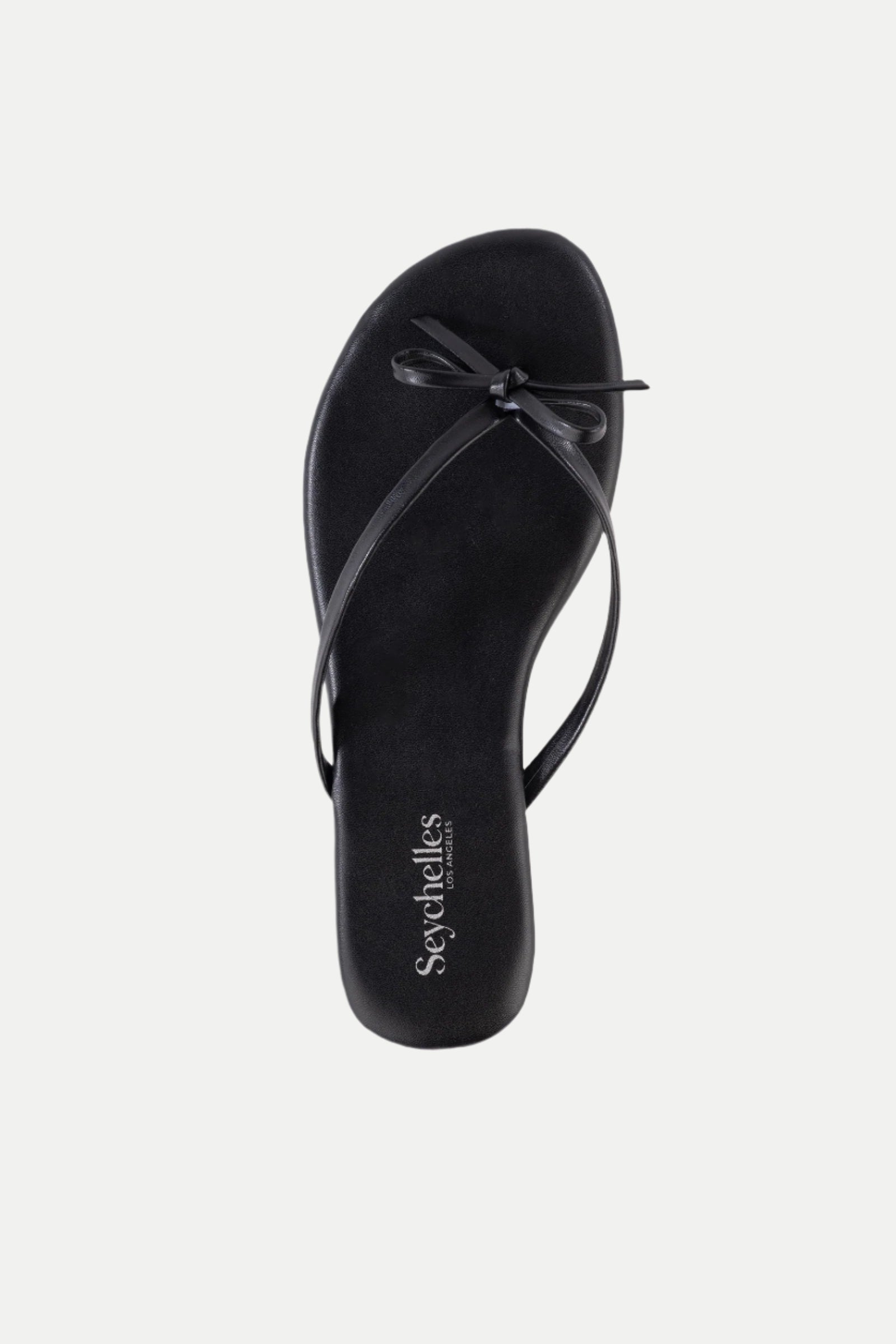 The Wish List Bow Thong Sandal is a black flat sandal with thin crisscross straps, a small bow on top, and Seychelles branding on the insole.