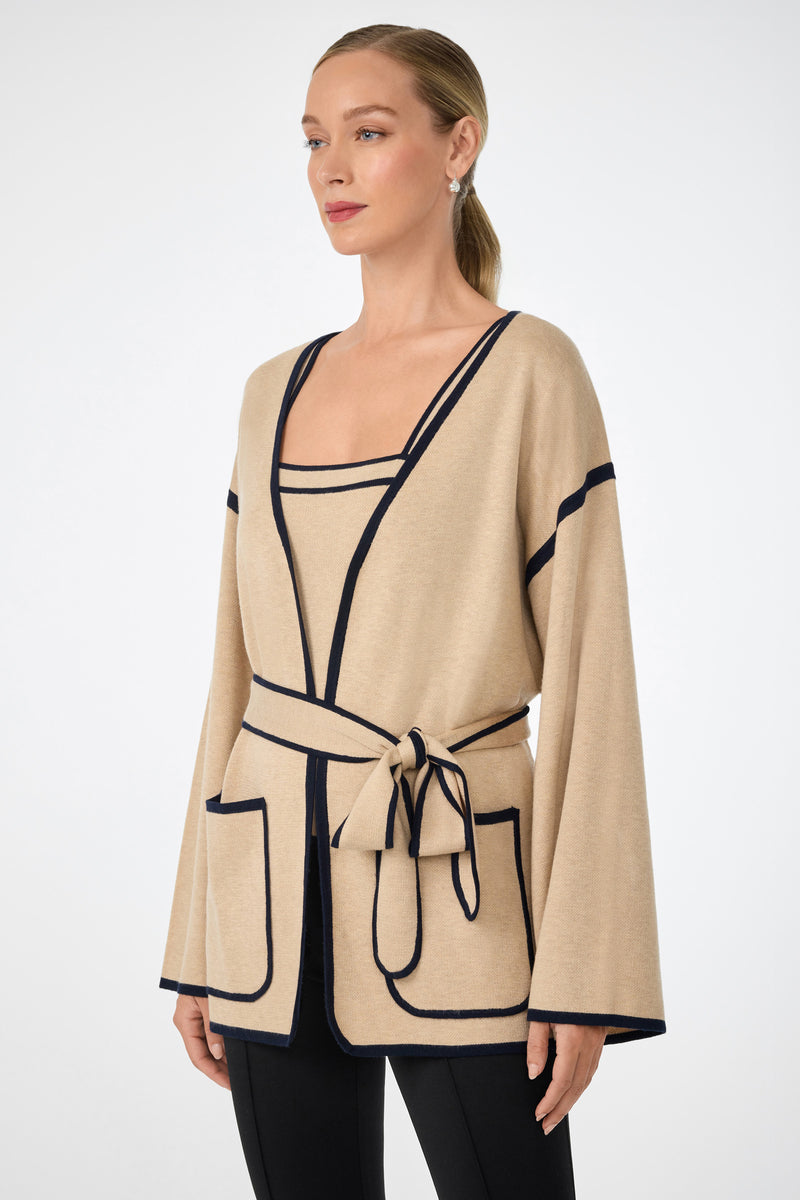 A woman wears the Wright Wrap Cardigan in beige with black trim and large pockets, layered over a matching square-neck top and paired with black pants, standing slightly turned against a plain light background.