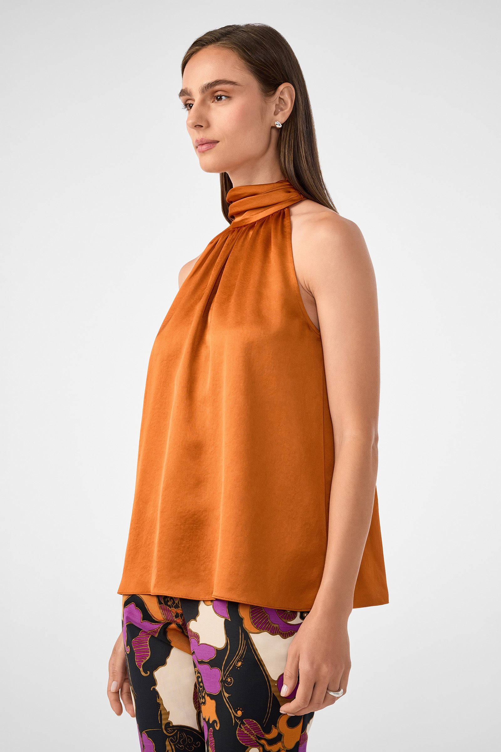 A woman in the Zana Top, a sleeveless high-neck orange satin blouse, and colorful abstract-patterned pants stands sideways against a plain light background.