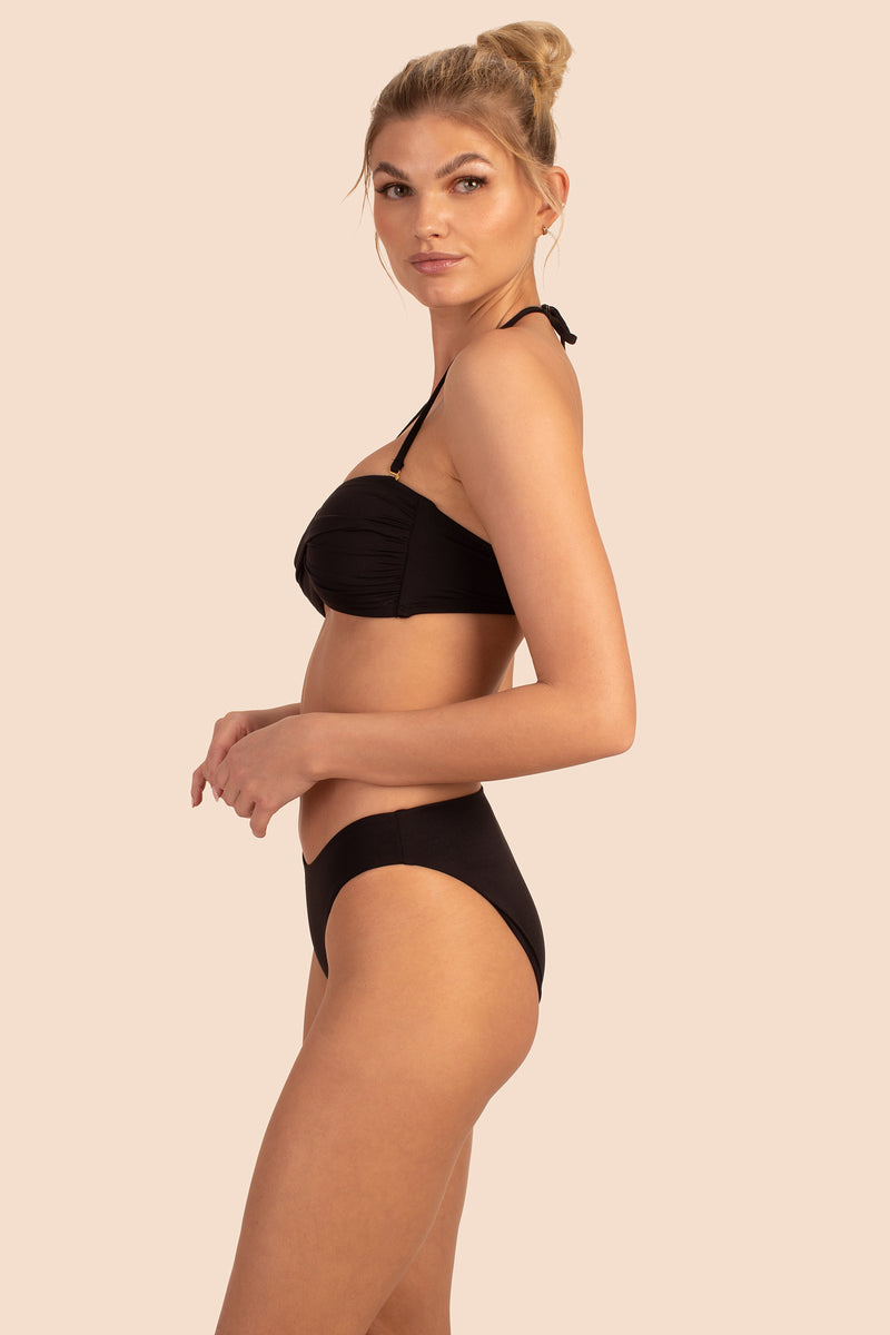 A blonde woman with her hair in a bun models the Women's Monaco Low Rise French Cut Swim Bottom in black, shown in profile against a plain beige background.