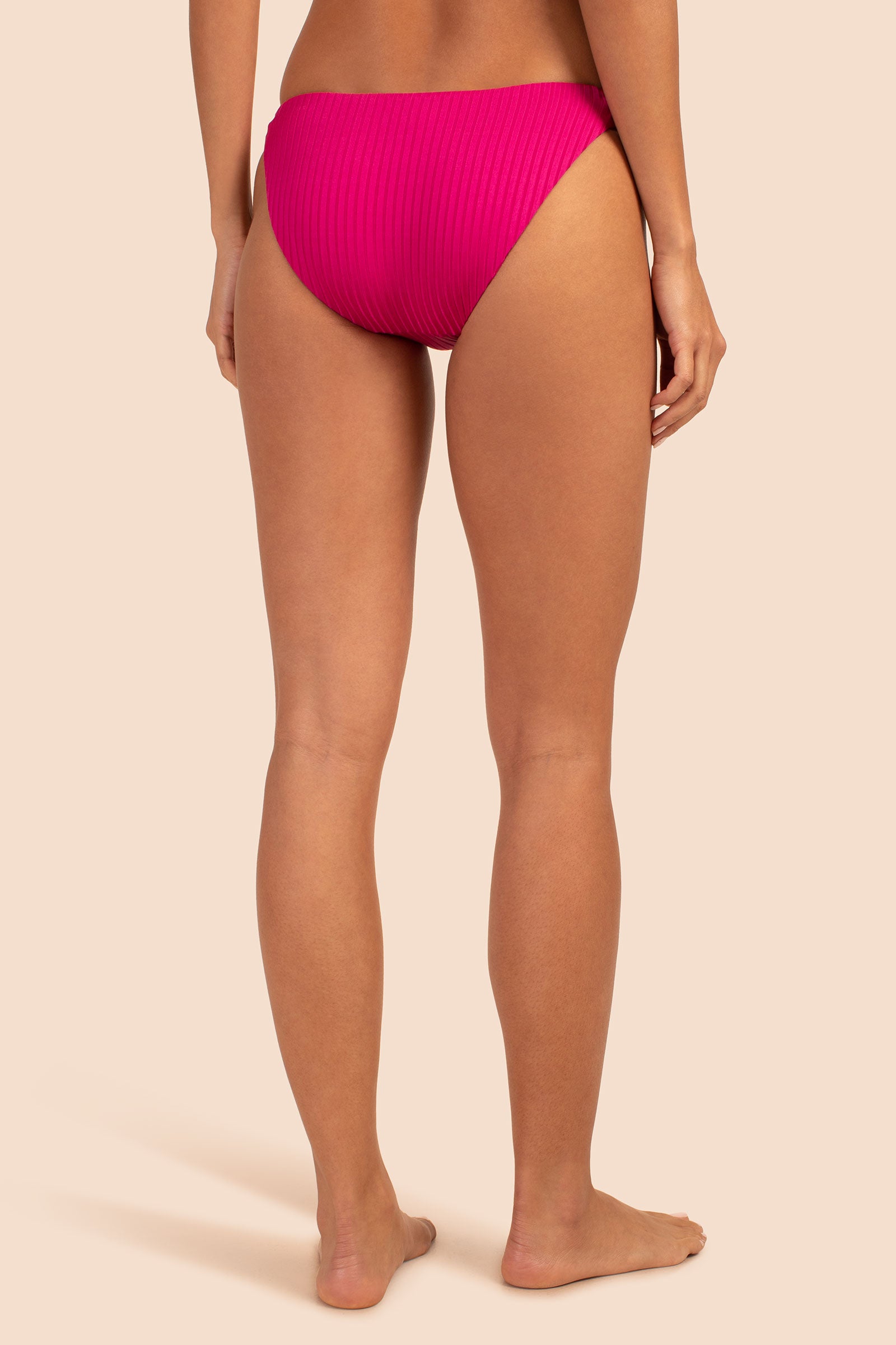 A woman stands barefoot facing away, modeling the Women's Olympia Rib Low Rise French Cut Swim Bottom in bright pink. Only her lower back, hips, and legs are visible against a plain light beige background.