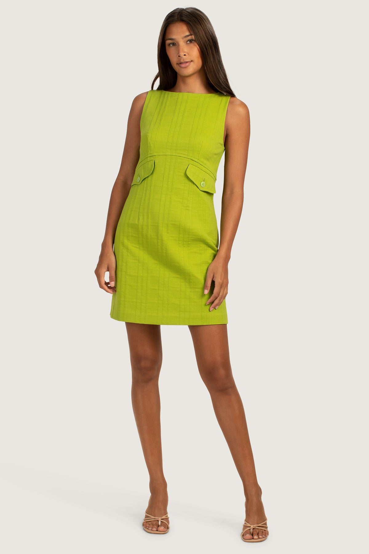 A woman models the Atrina Dress, a sleeveless lime green boatneck sheath with check weave jacquard and two front flap pockets. She stands confidently, with long straight hair and clear heeled sandals.