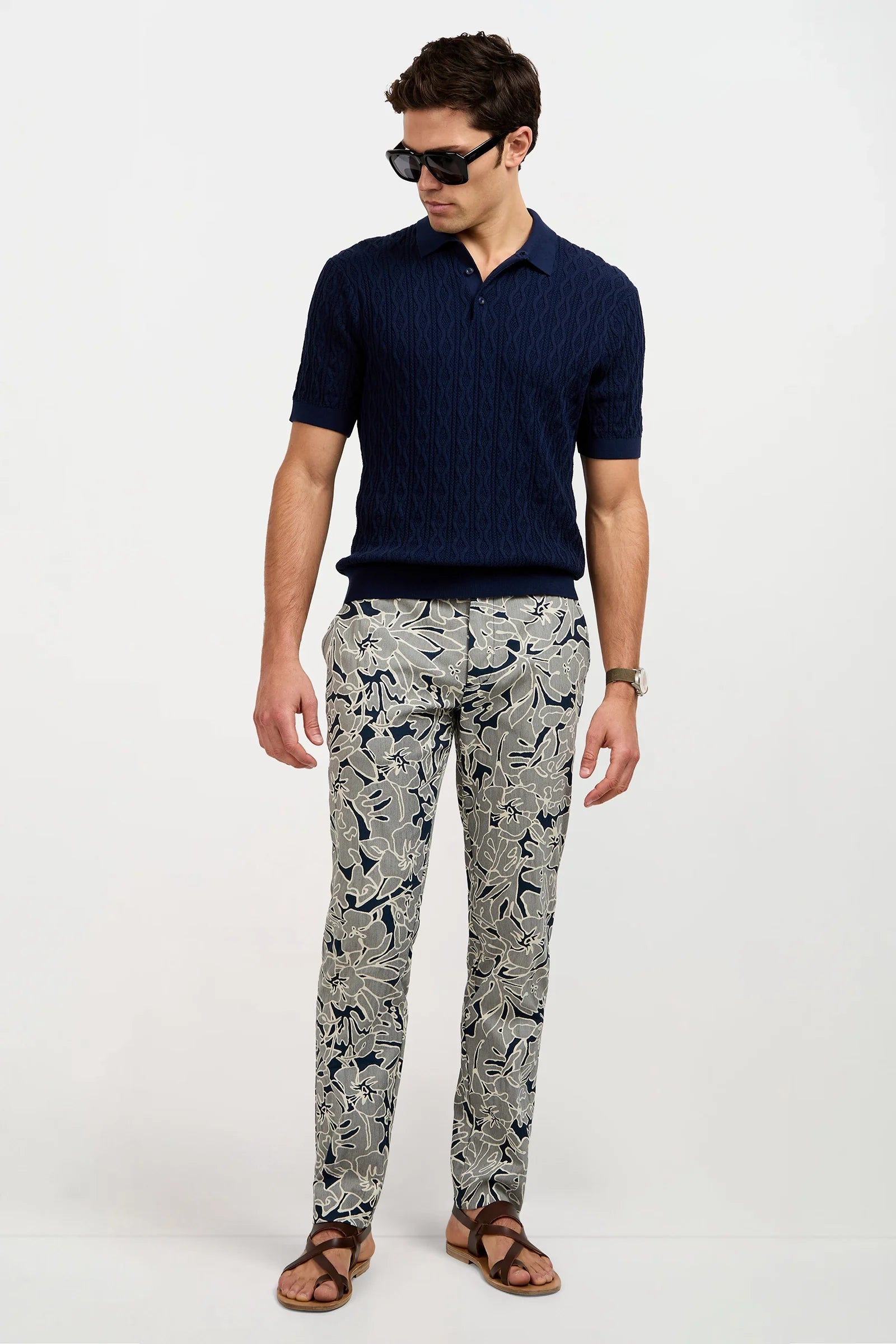 A man wearing sunglasses, a navy short-sleeved polo shirt, patterned beige and navy trousers with a leafy design, brown sandals, and a watch, stands against a plain white background.
