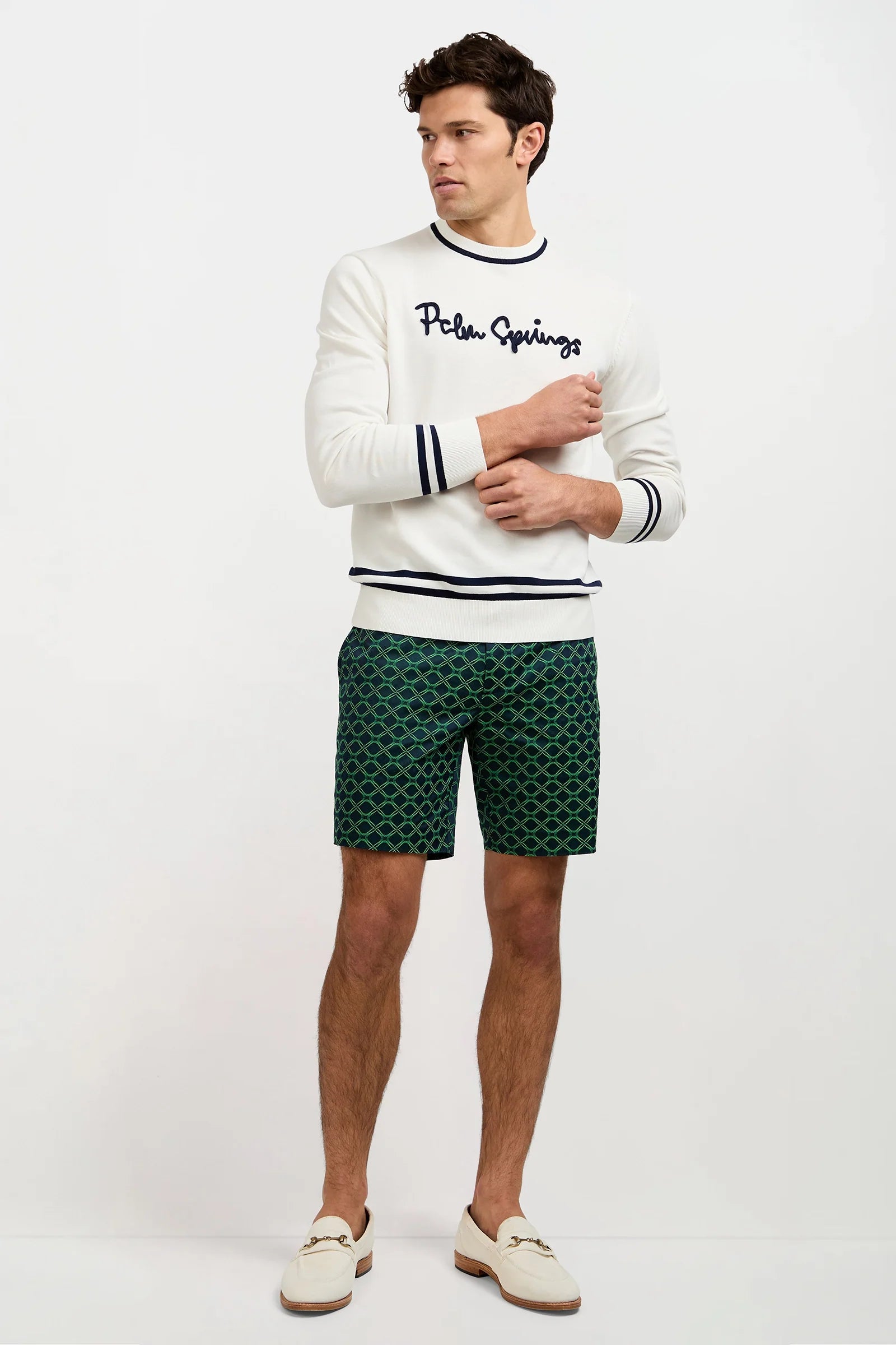 A man stands wearing a white sweater with Palm Springs written on it, green patterned shorts, and cream loafers. He looks to the side with his arms crossed in front of a plain white background.