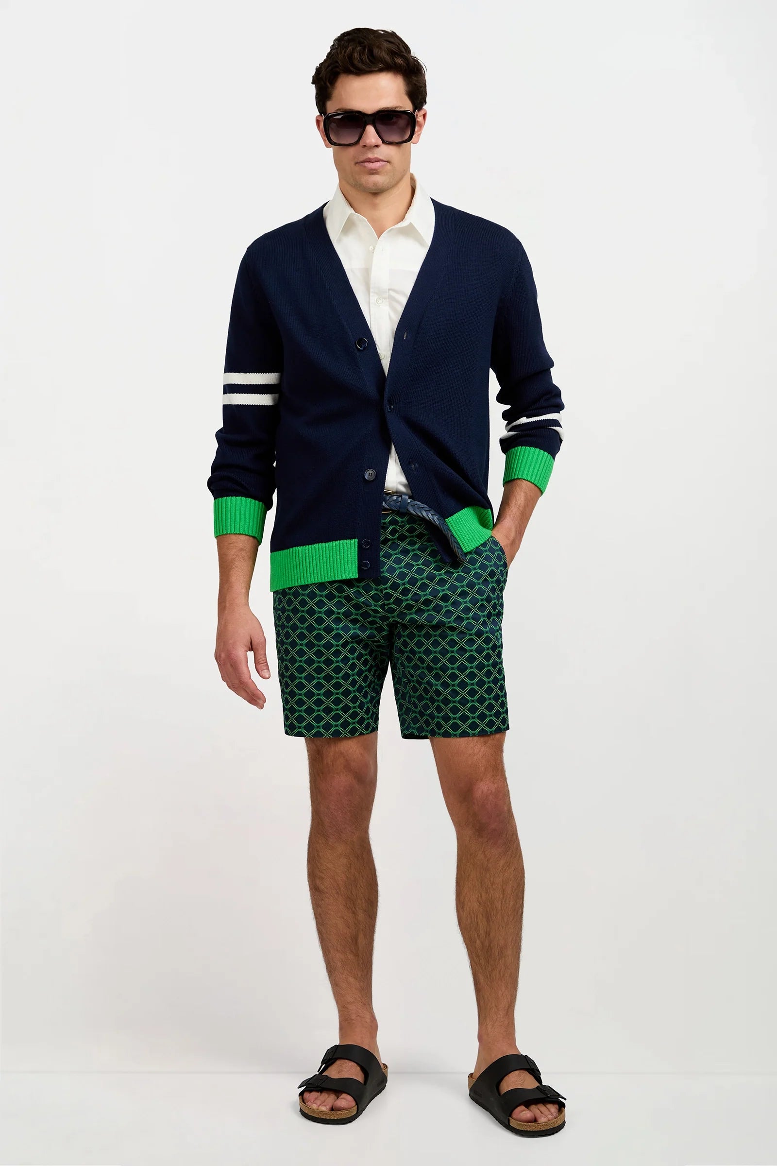 A man wearing dark sunglasses, a navy cardigan with green cuffs, a white shirt, patterned green shorts, and black sandals stands against a plain white background.
