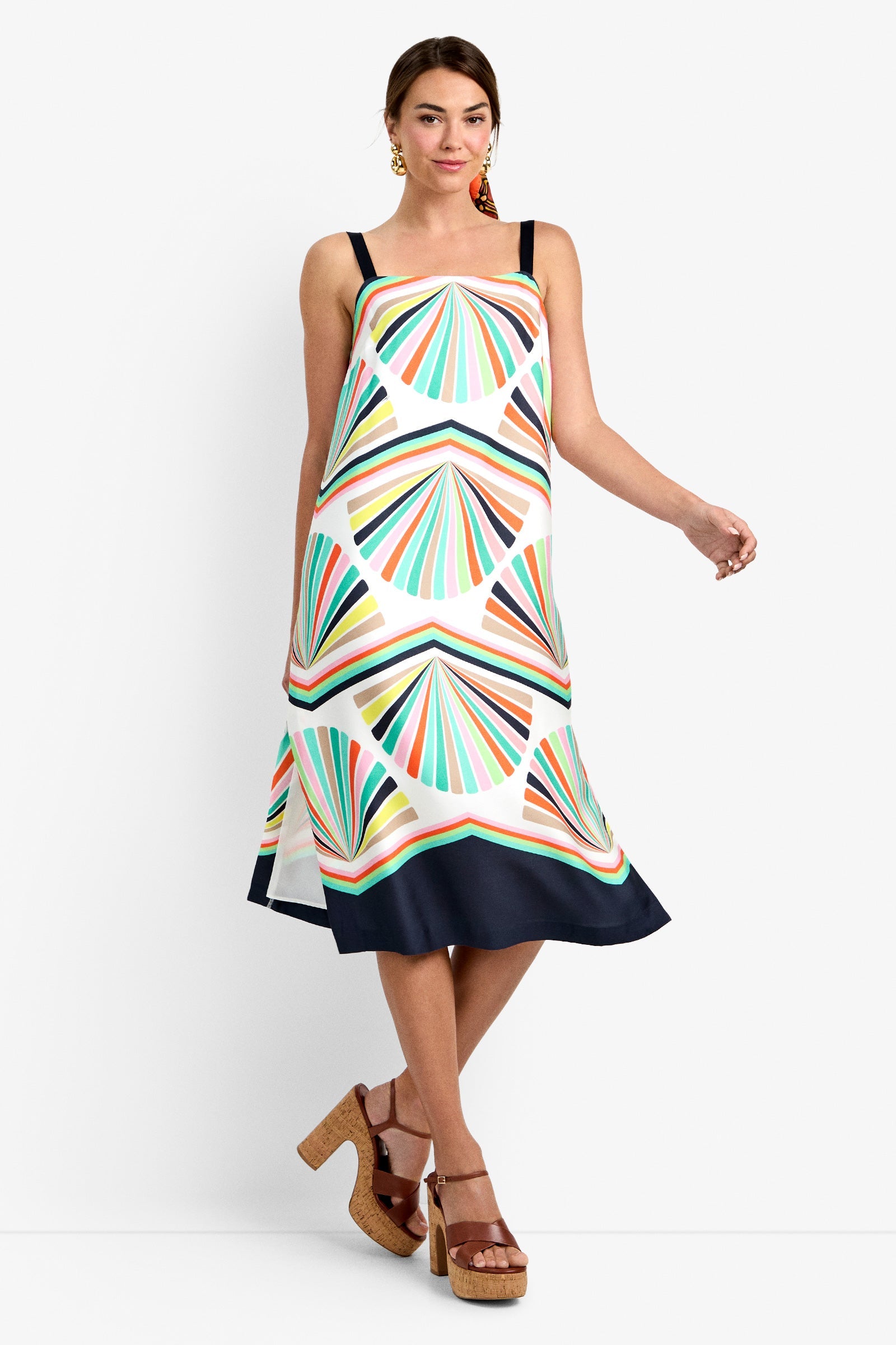 A woman wearing a sleeveless, geometric-patterned midi dress with colorful fan shapes and a dark hem, paired with brown platform sandals, poses against a white background.
