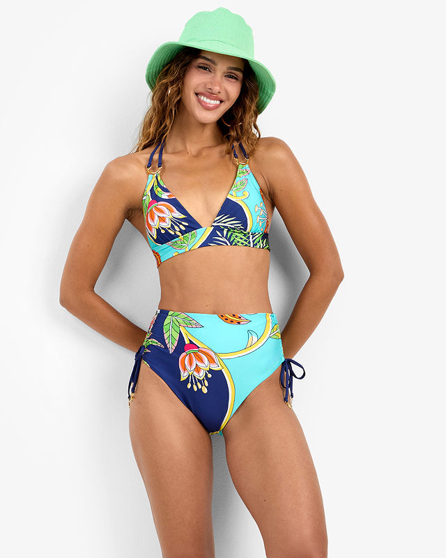 Woman wearing a large, striped sunhat and a halter top leans on a white railing by the sea, with blue sky and water in the background. She wears white pants and green earrings, looking thoughtfully into the distance.
