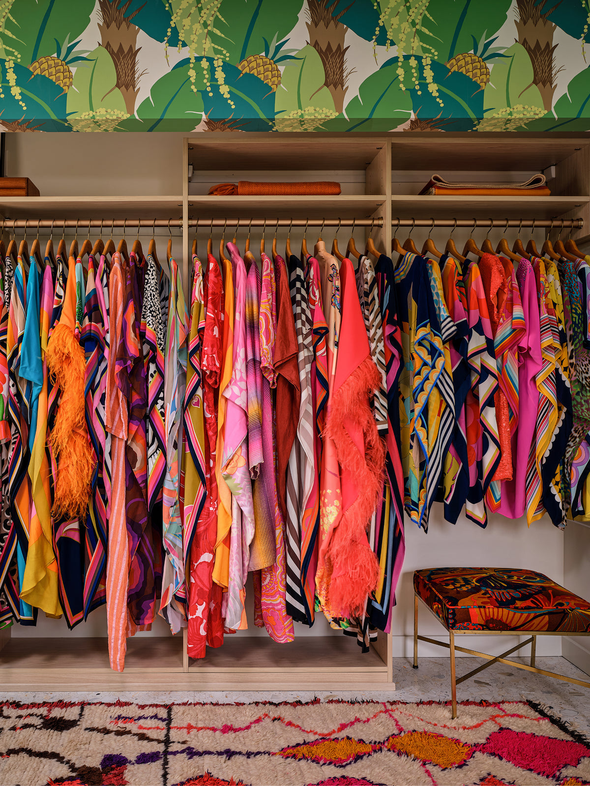 A colorful closet with vibrant clothing, including dresses and robes, hanging neatly on wooden hangers. Tropical wallpaper and a patterned bench complete the lively, eclectic space.