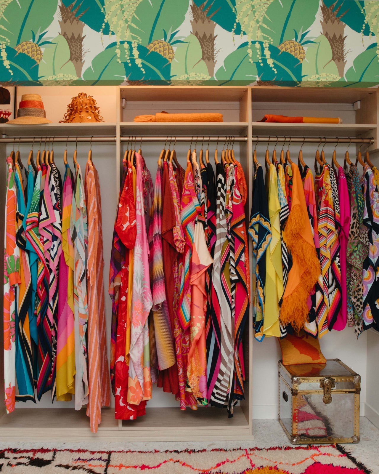 A colorful closet with vibrant clothing, including dresses and robes, hanging neatly on wooden hangers. Tropical wallpaper and a patterned bench complete the lively, eclectic space.