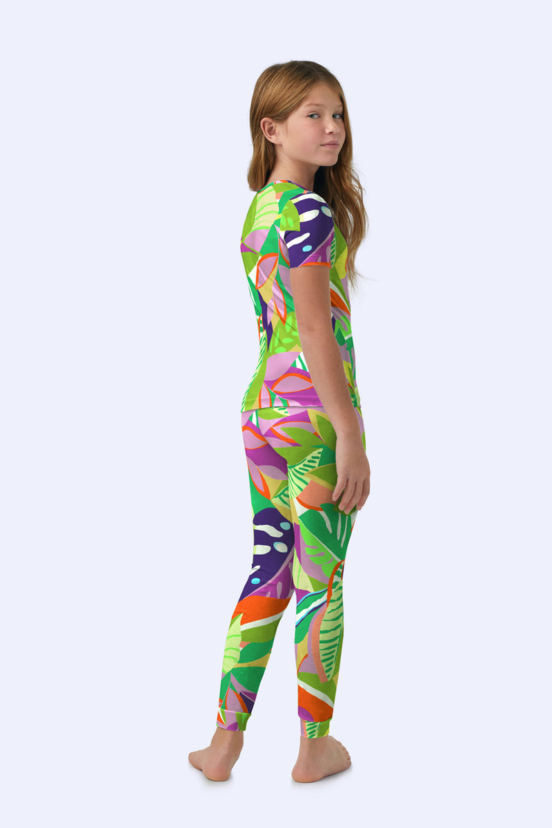 A young girl with long brown hair stands barefoot, slightly turned to the side, wearing the Kids PJ Set - Jungle Stretch Jersey featuring a colorful tropical leaf pattern in green, pink, orange, and purple on a light background.