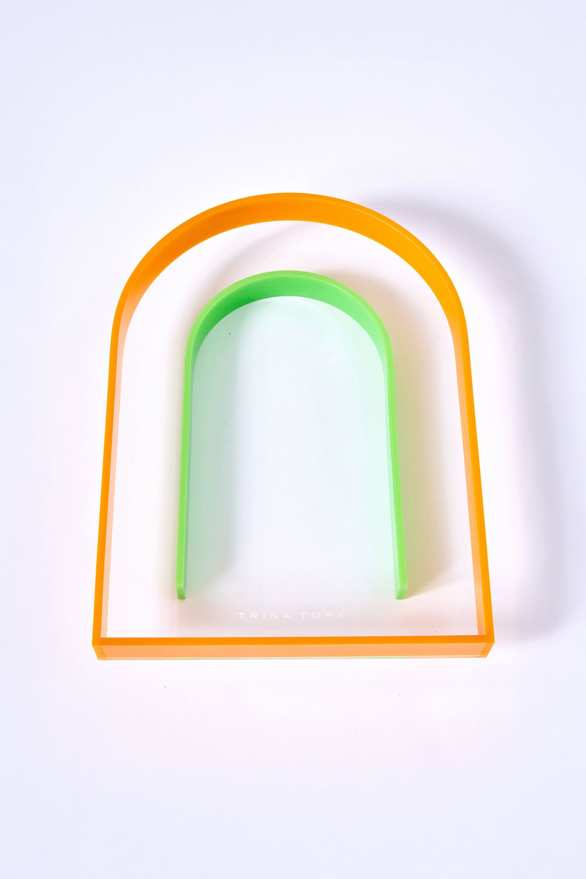 The Arched Acrylic Serving Tray is a clear, rectangular tray with rounded top edges, an orange border, and a green U-shaped curve inside. Trina Turk appears subtly near the bottom center.