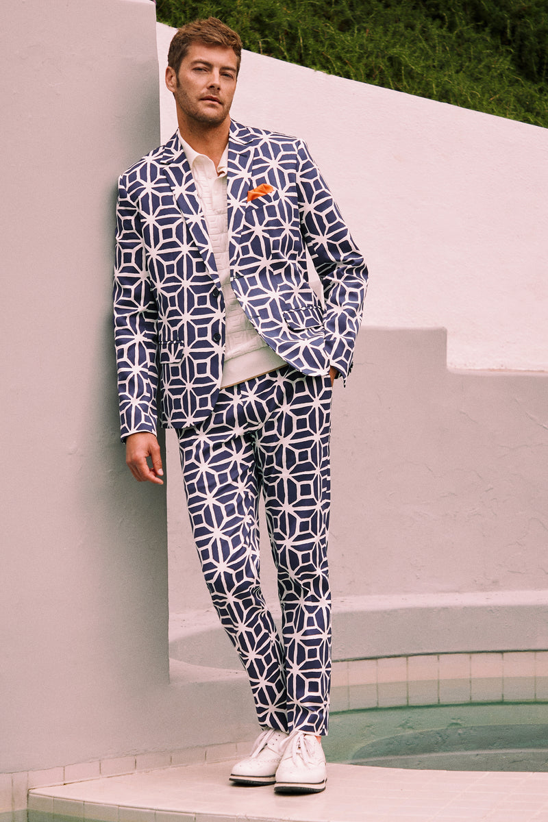 A man stands by a pool in Clyde Slim Trouser - Durell Herringbone, styled with a white shirt, white sneakers, an orange pocket square, against white walls and green grass.