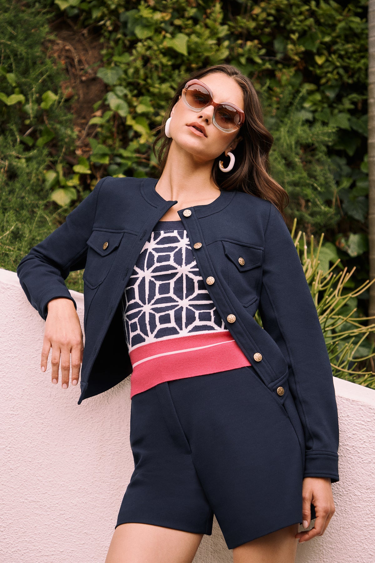 A woman poses outdoors against a light pink wall, wearing sunglasses, large hoop earrings, a navy jacket, a geometric-patterned top, and the Valera Shorts - Stretch Piqué. Green foliage appears in the background.