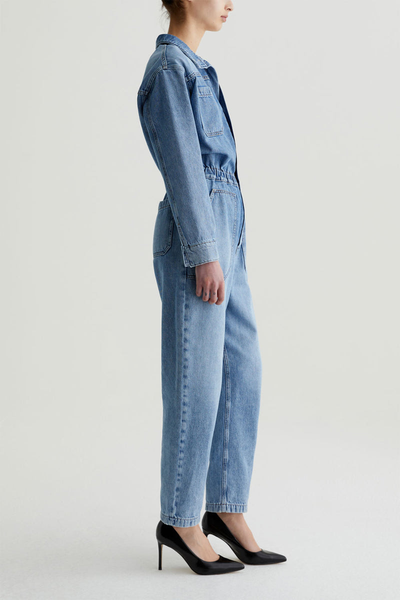 A woman stands sideways in the AG Nita Denim Jumpsuit with long sleeves and chest pockets, paired with black heels, against a plain white background.