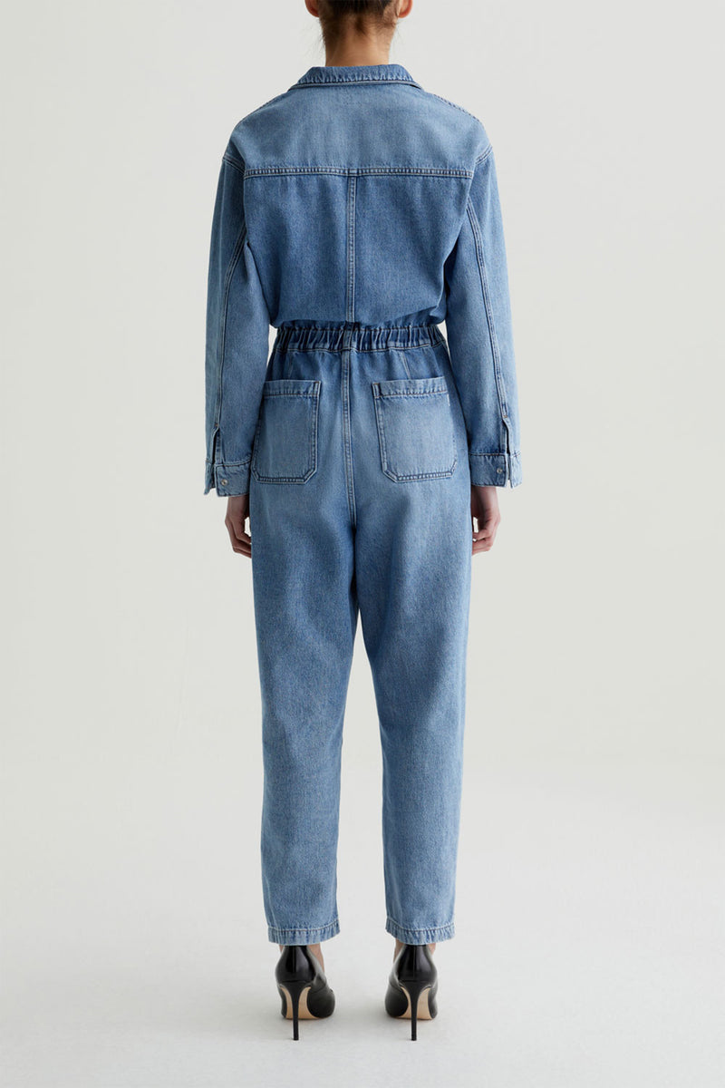A person stands facing away, wearing the AG Nita Denim Jumpsuit with long sleeves and an elastic waistband, paired with black high heels. The background is plain and light-colored.