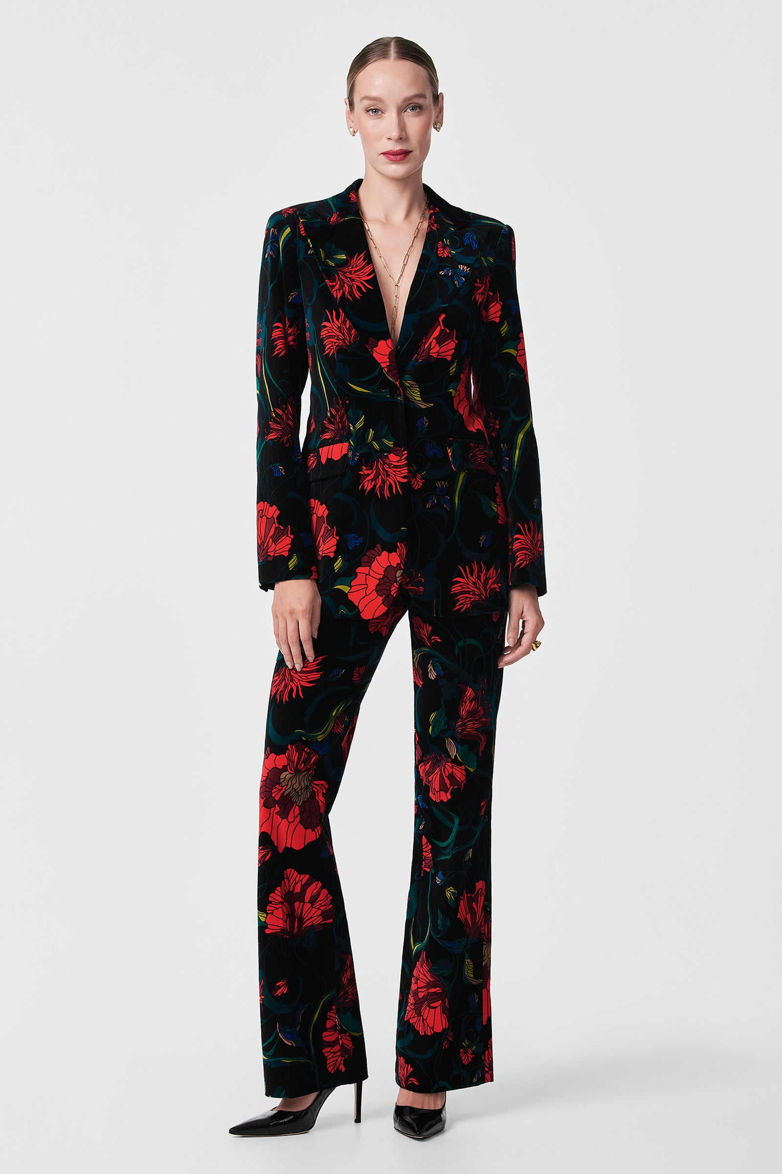 A woman with light skin and pulled-back hair wears the Broadway Printed Velvet Jacket, featuring a bold red floral pattern, paired with black pointed heels. She stands against a plain background and sports minimal jewelry.