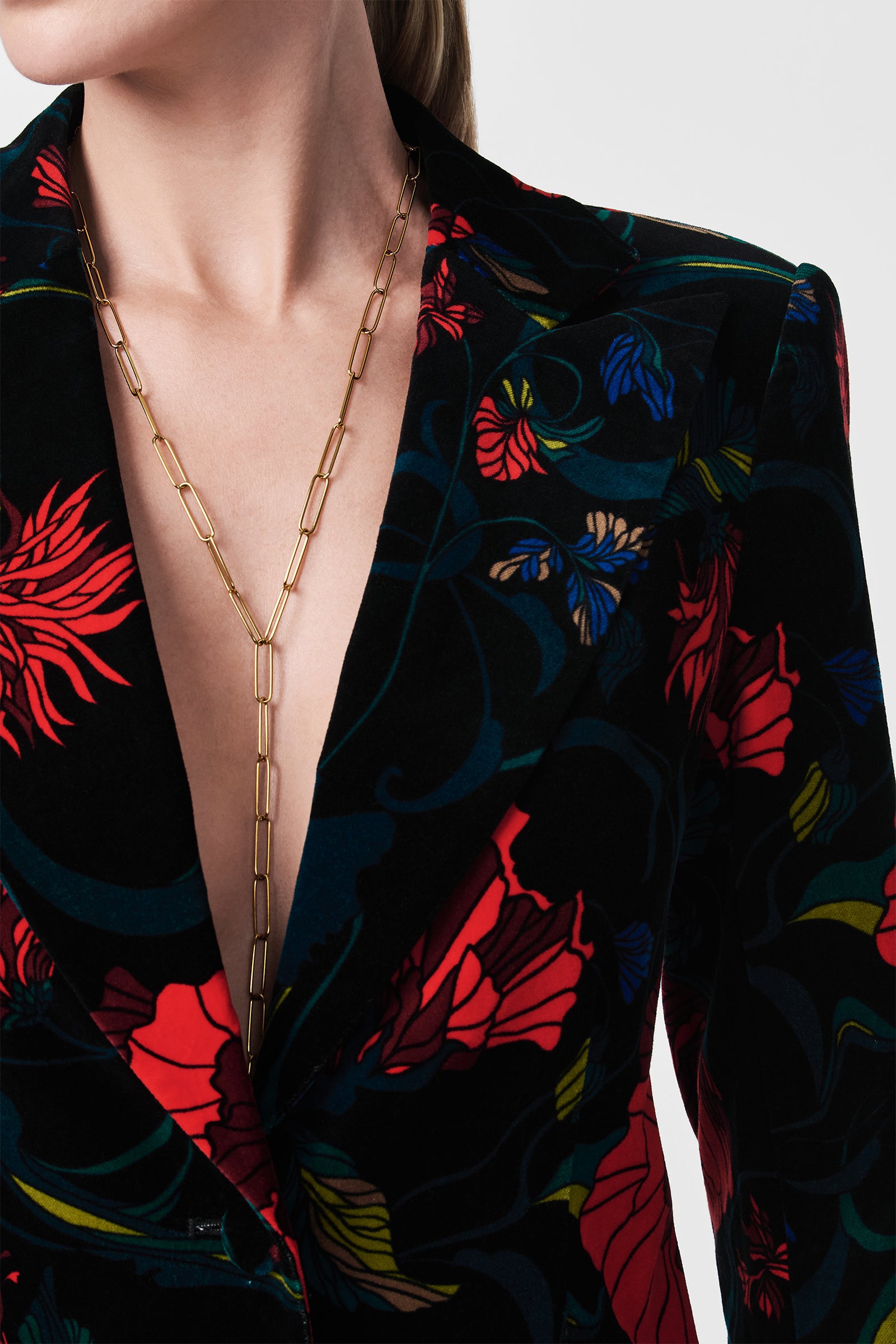 A person wears the Broadway Printed Velvet Jacket—a black blazer with red, blue, and yellow floral pattern and a deep V-neck—paired with a gold chain necklace. The background is light and plain.