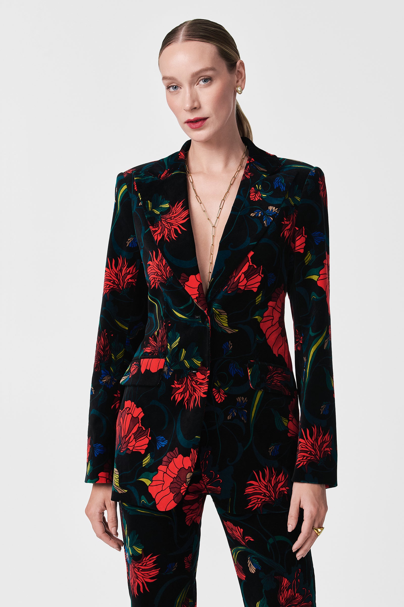 A woman with slicked-back hair wears the Broadway Printed Velvet Jacket, featuring bold red, blue, and green floral patterns and a deep V-neckline, paired with a gold chain necklace against a plain white background.
