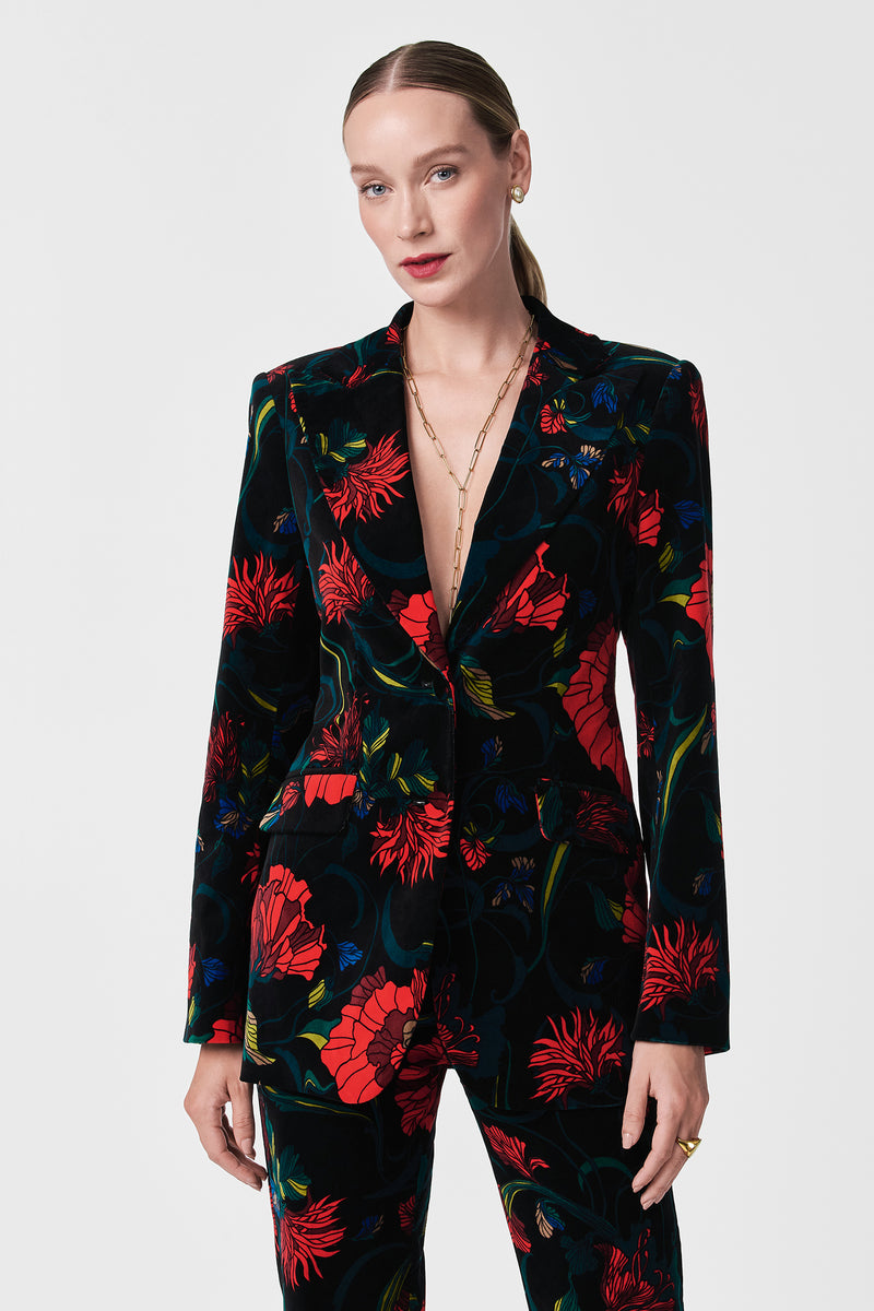 A woman with slicked-back hair wears the Broadway Printed Velvet Jacket, featuring bold red, blue, and green floral patterns and a deep V-neckline, paired with a gold chain necklace against a plain white background.