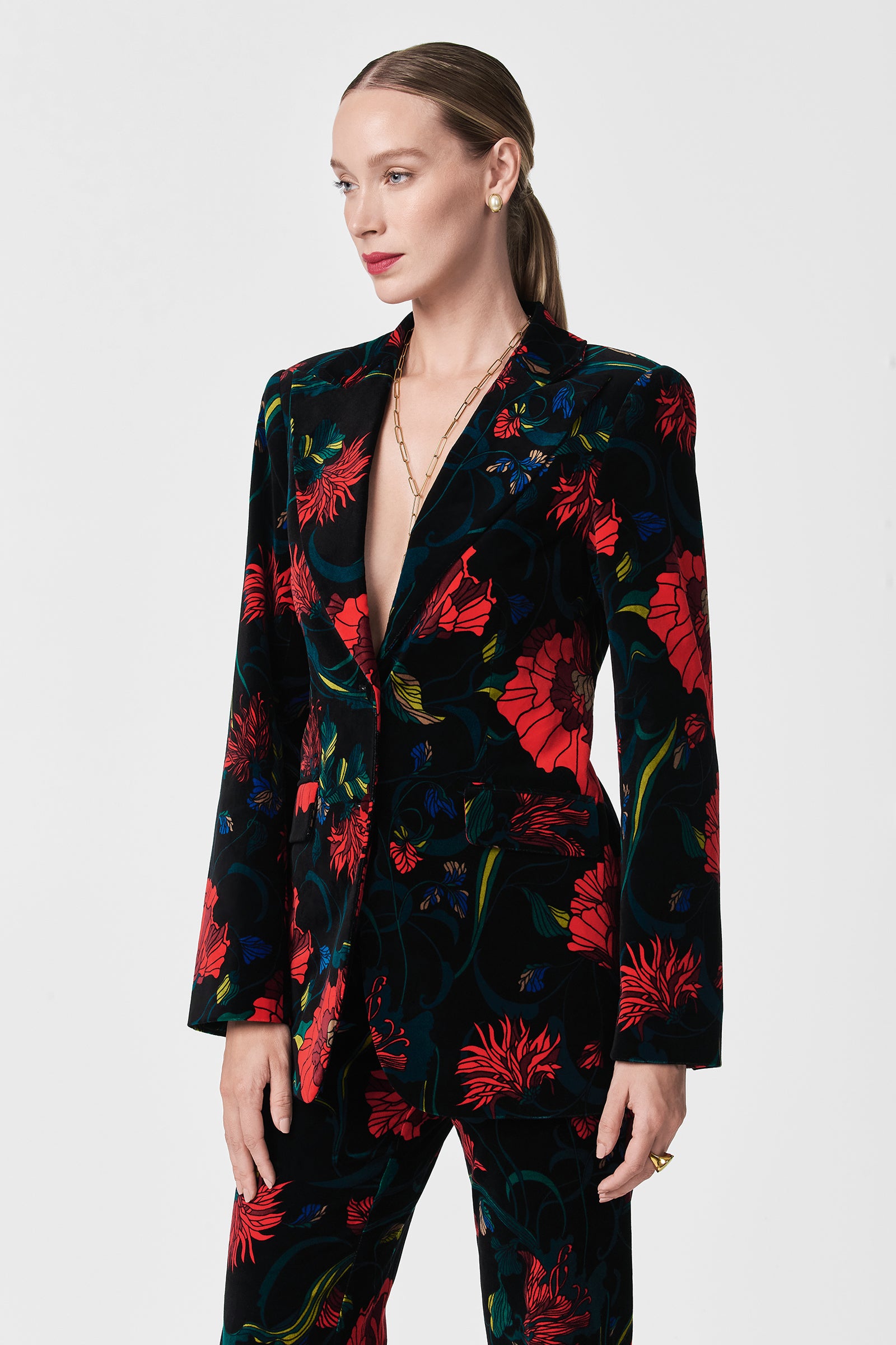 A woman with slicked-back hair confidently models the Broadway Printed Velvet Jacket, featuring a striking red and green floral pattern on black velvet, while standing against a plain white background.