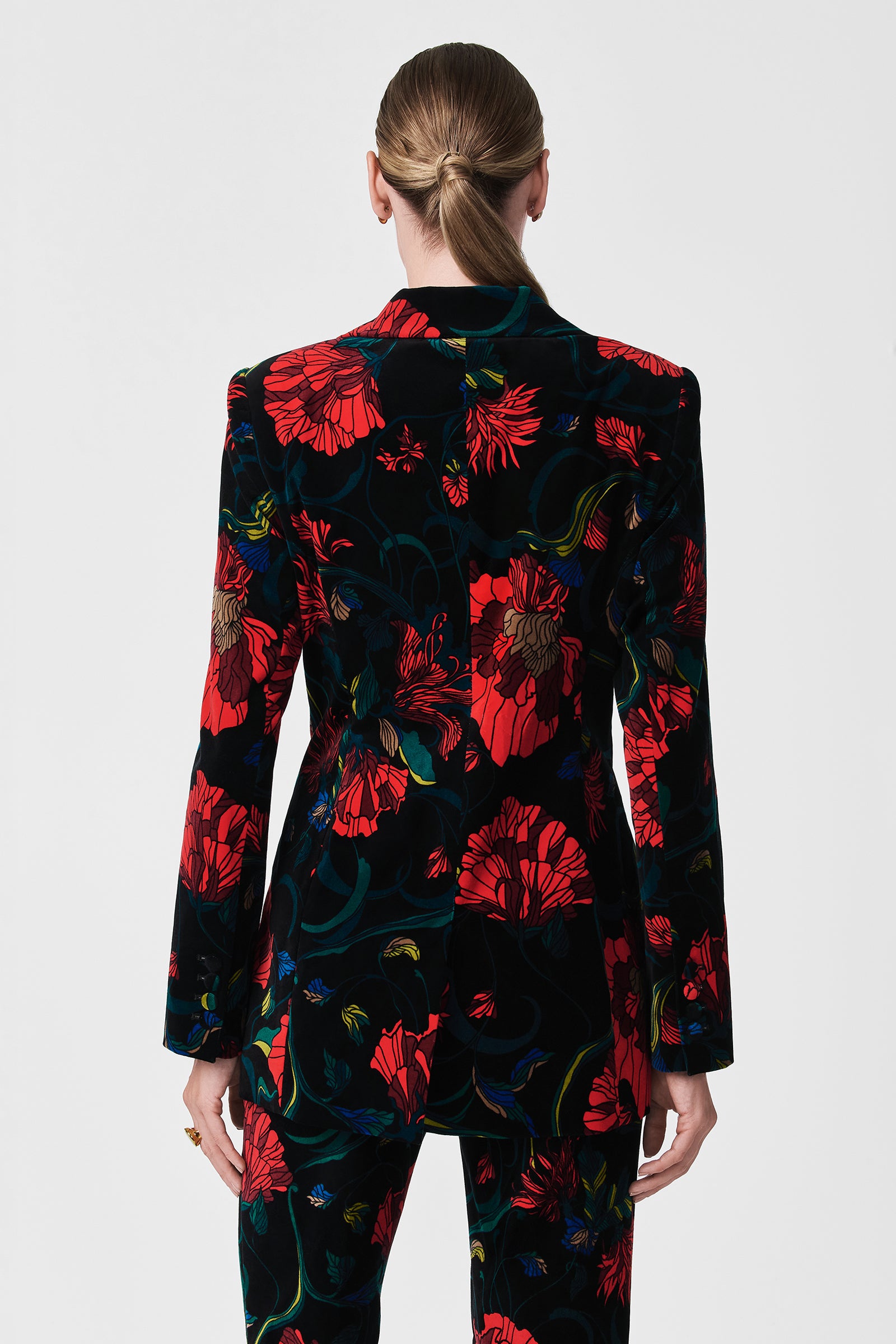 A person with blonde hair in a low bun is shown from the back wearing the Broadway Printed Velvet Jacket, featuring a bold red and green floral pattern, against a plain white background.