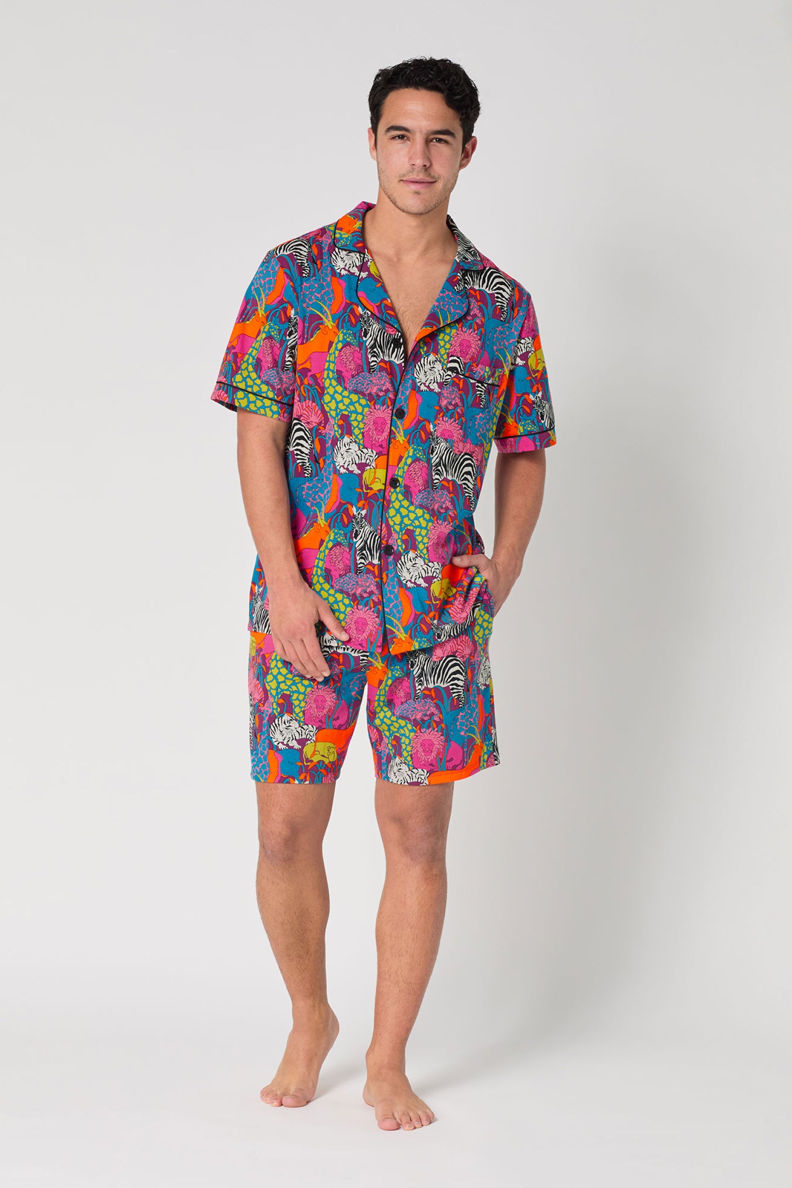 A man stands barefoot wearing the Men's Boxer PJ Set - Jungle in MULTI, featuring a vibrant animal and floral pattern, short-sleeved button-up shirt, and matching shorts on a plain white background.