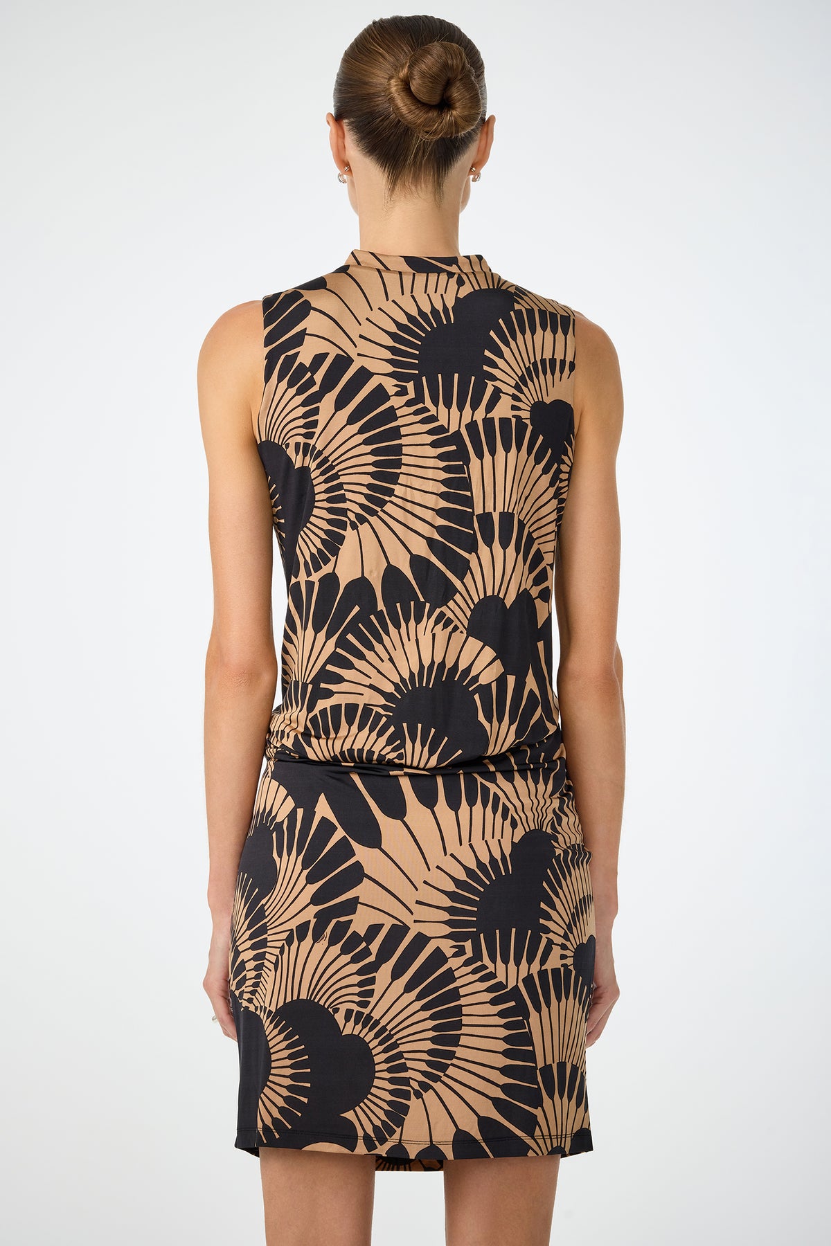 A woman with her hair in a neat bun is seen from the back wearing the Lenaya 3 Drape Dress - Juniper Jersey, a sleeveless, knee-length dress featuring a tan and black abstract fan pattern, set against a plain light background.